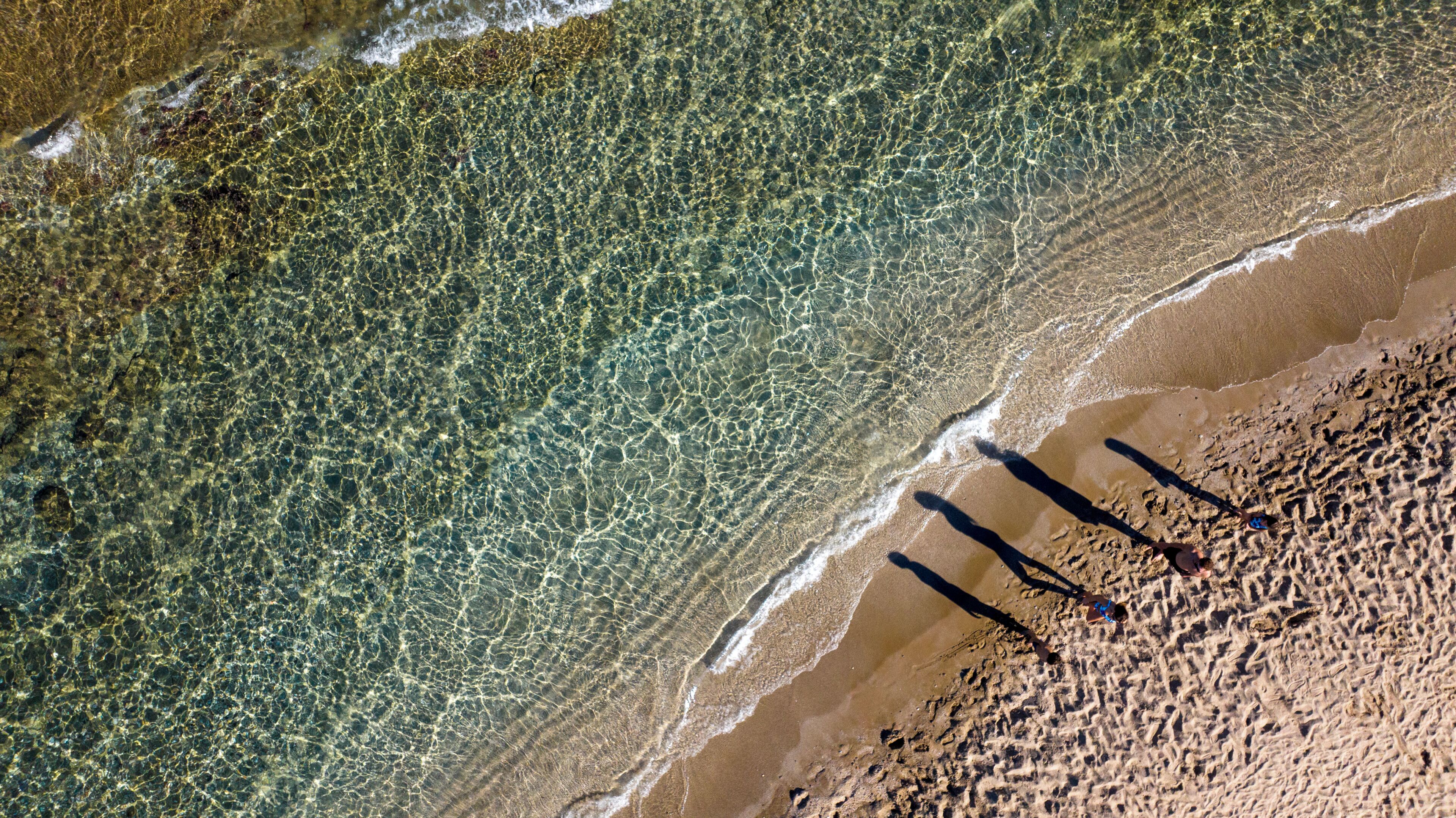 Vista aerea di una spiaggia con acqua cristallina, disegni di sabbia ben visibili e ombre di persone.