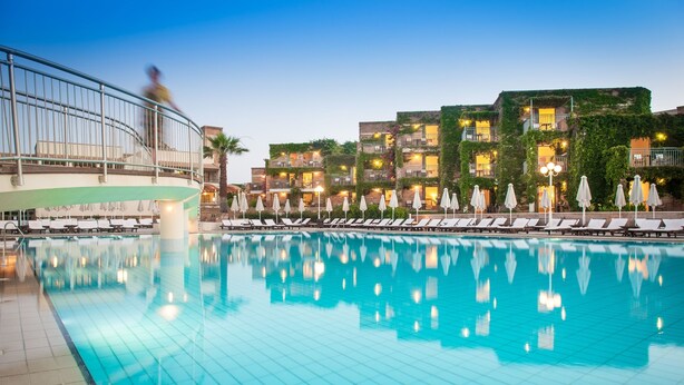 Una piscina con sillas de descanso y sombrillas, rodeada de edificios con balcones y luces.