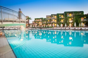 A swimming pool with lounge chairs and umbrellas, surrounded by buildings with balconies and lights. - Bella Beach - All Inclusive (Hersonissos)