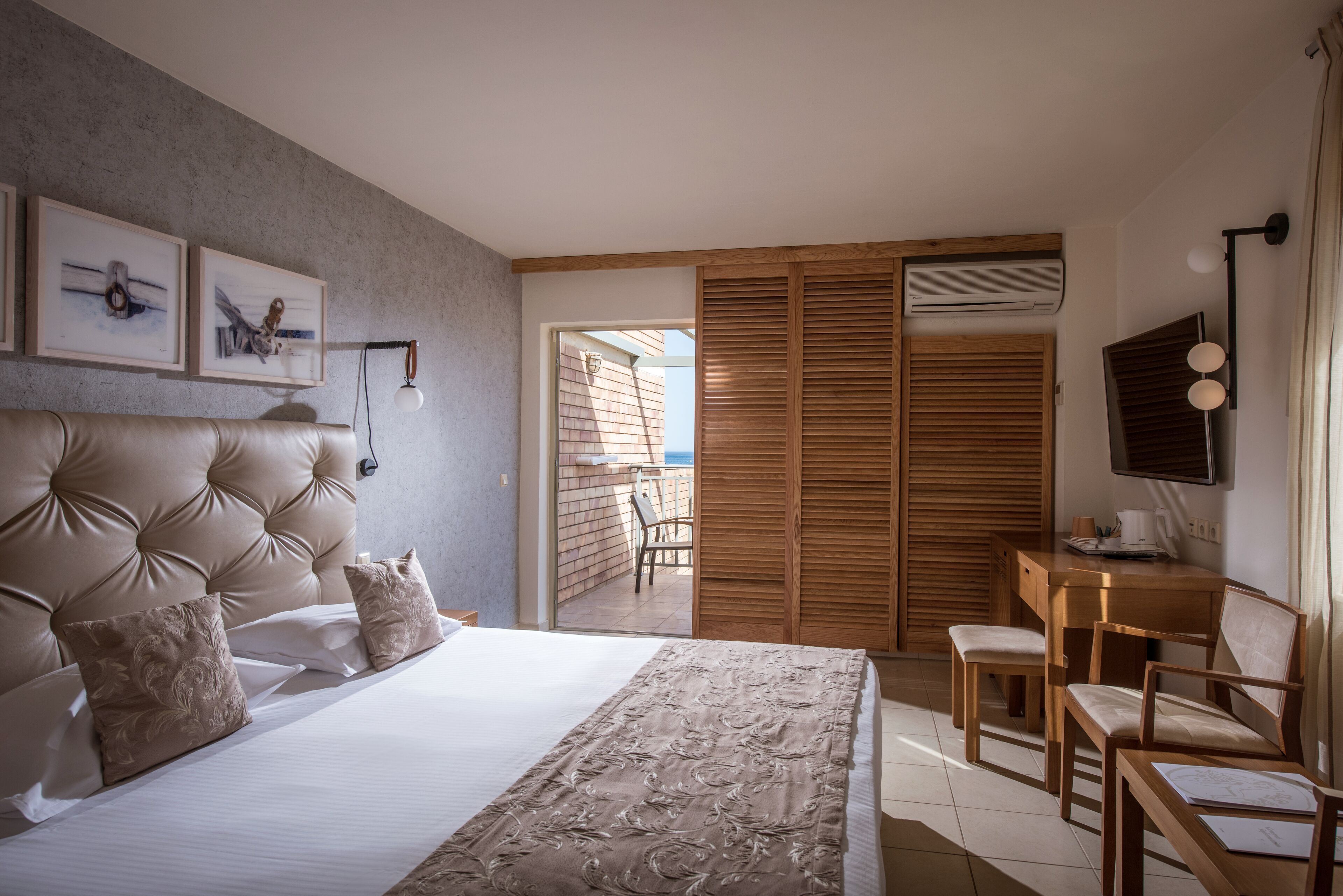 A bedroom with a large bed, a wooden desk, and a TV. There is a balcony with a chair and a table.