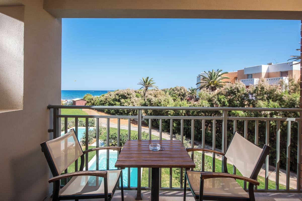 Ein Balkon mit Tisch und Stühlen, der auf einen Pool und den Blick auf Meer und Gebäude bietet.