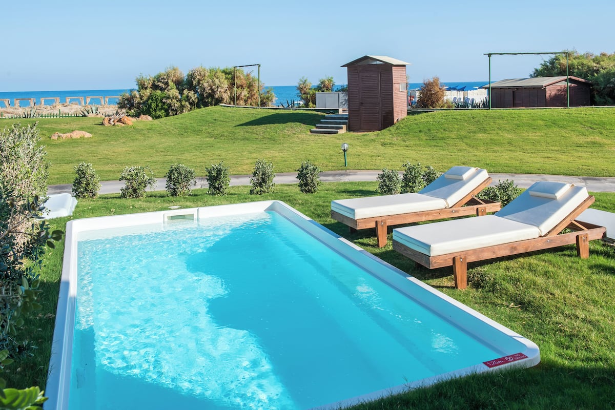 Ein Schwimmbecken mit zwei Liegestühlen und einer kleinen Gartenhütte in einer grünen Umgebung mit Blick auf das Meer.
