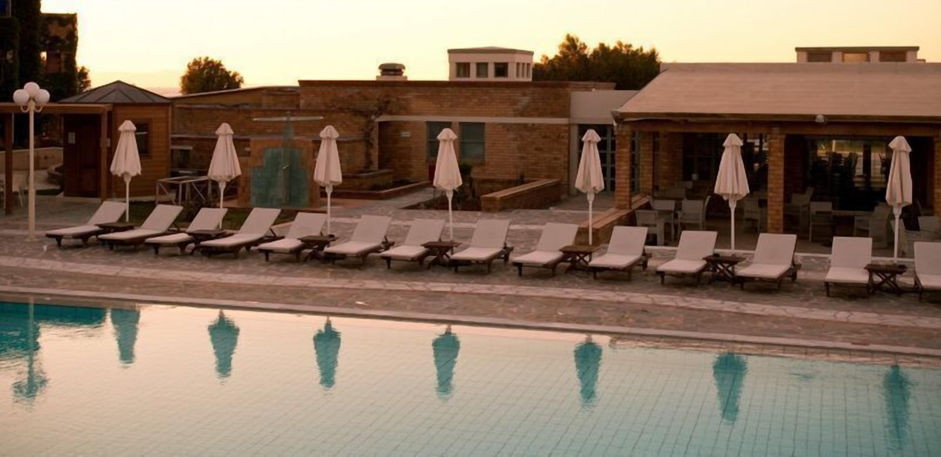 A poolside area with lounge chairs and umbrellas, a tiled pool, and buildings in the background.