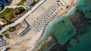 Vista aérea de una playa con filas de sombrillas y tumbonas, un estacionamiento y un edificio con techo curvo.