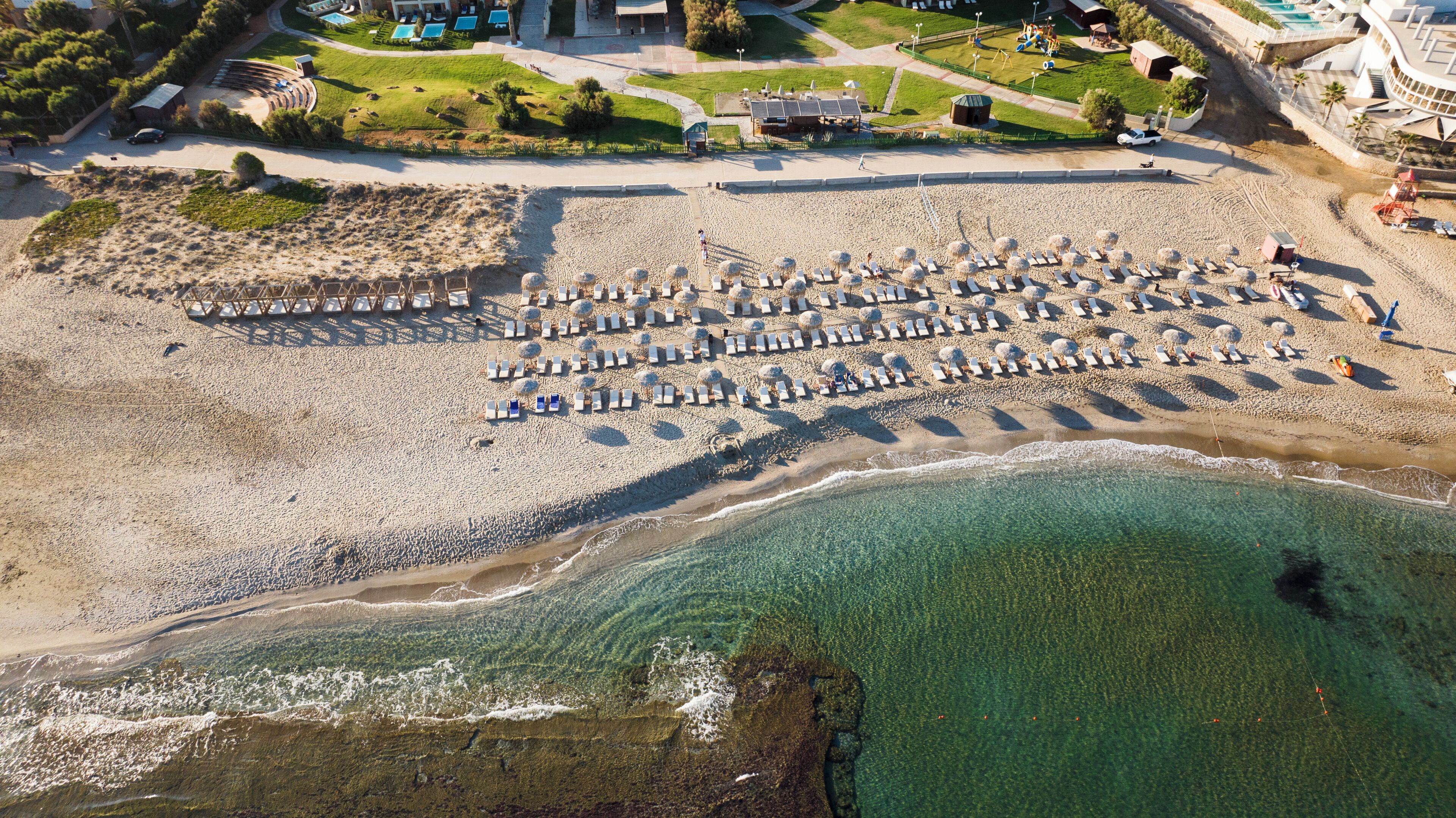 Une vue aérienne d’une plage avec des rangées de transats et de parasols, une mer bleue et limpide, et un bâtiment avec une piscine en arrière-plan.