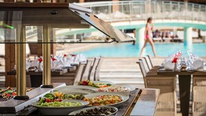 Una mesa de buffet con distintos platos, un área de piscina con tumbonas, y un puente al fondo.