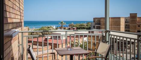 A balcony with a wooden table and two chairs, overlooking a beach with buildings in the background.