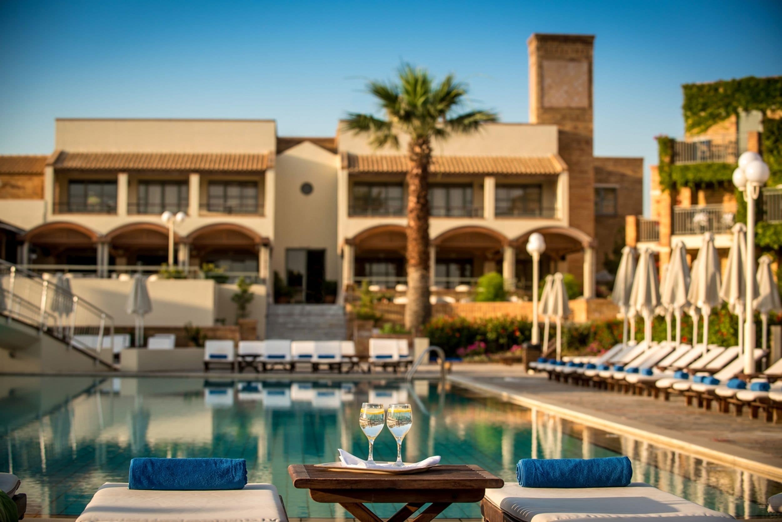 A poolside area with lounge chairs, a table with two glasses of water, and a building with multiple balconies and arches in the background.