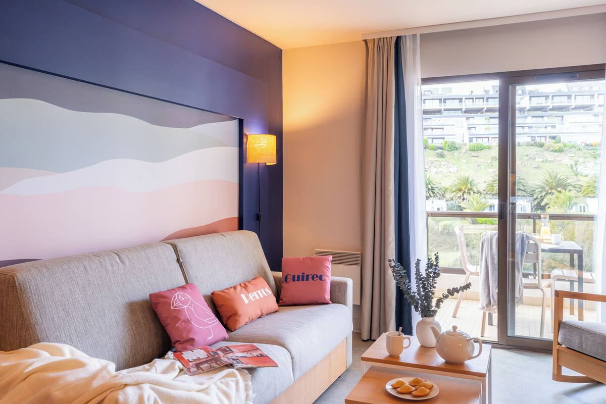 A modern hotel room with a sofa, pillows, a table with a teapot, and a view of the outdoor balcony.