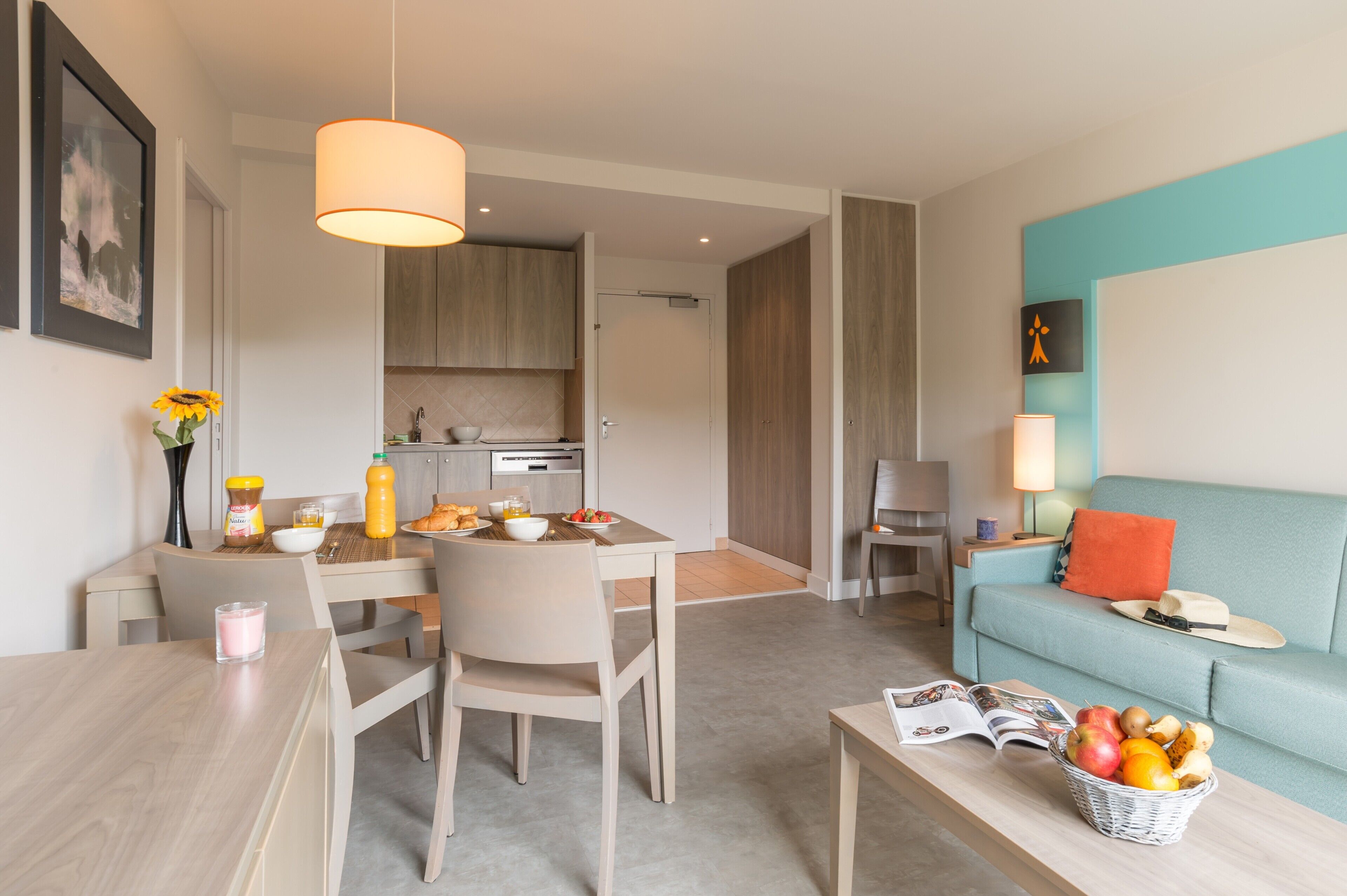 Een moderne woonkamer met een bank, een eettafel met stoelen, een keuken op de achtergrond en een fruitmand op een bijzettafeltje.