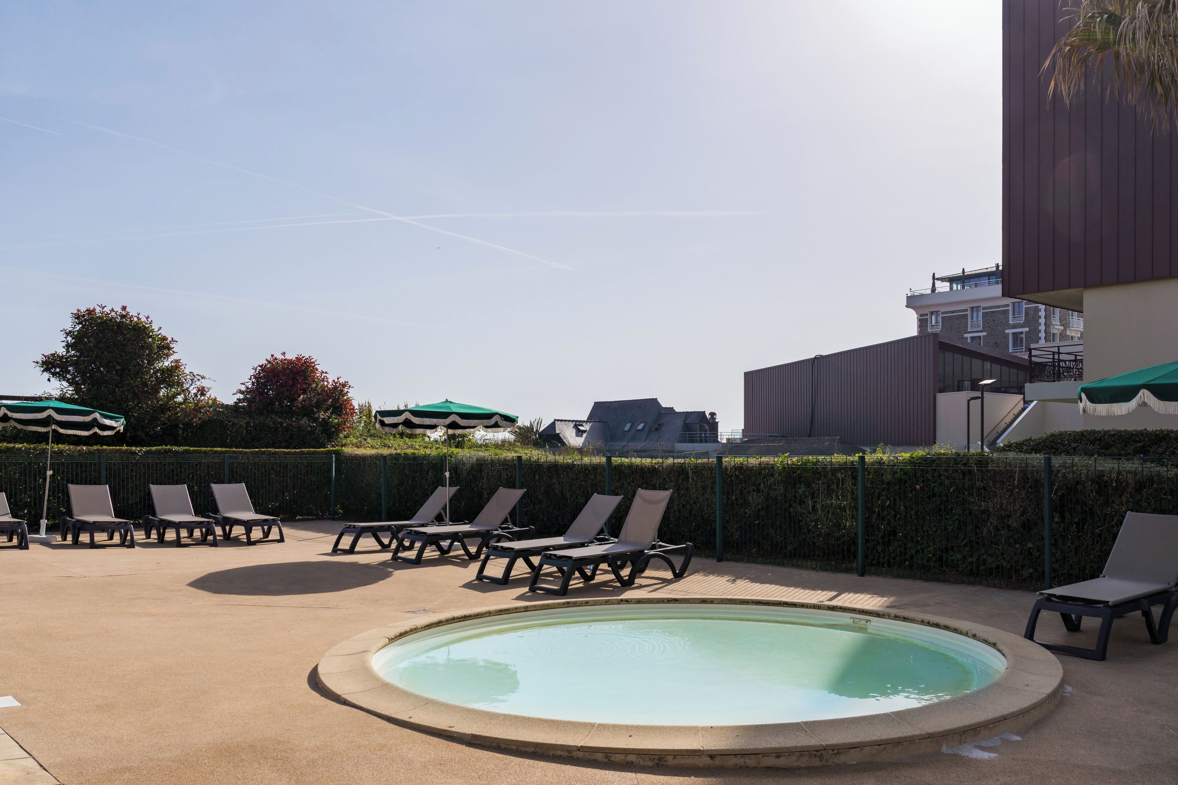 Un espace piscine avec des chaises longues et des parasols, entouré d’une haie et bordé de bâtiments en arrière-plan.