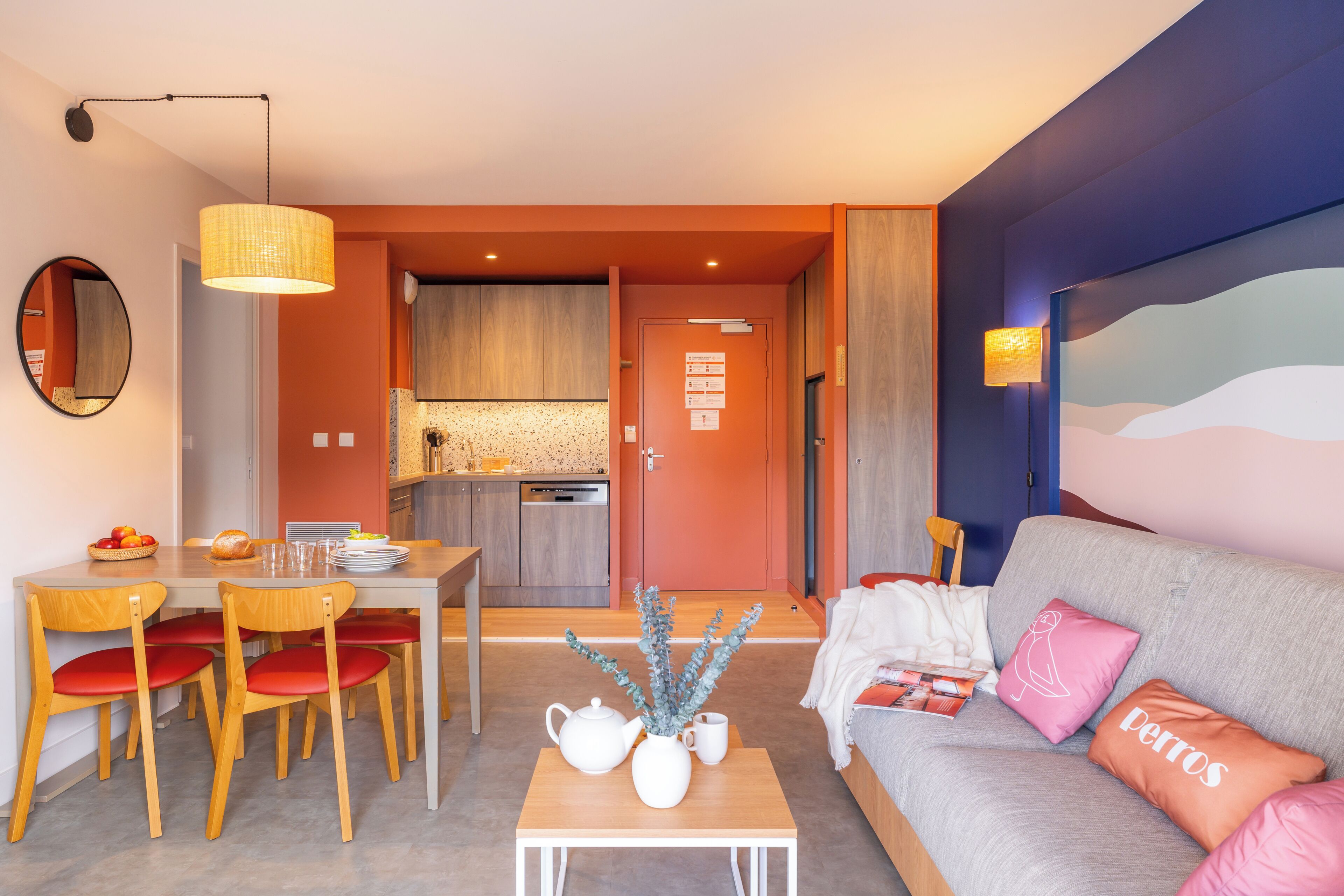 A modern living room with a sofa, a dining table with chairs, a kitchen in the background, and a large wall mural.