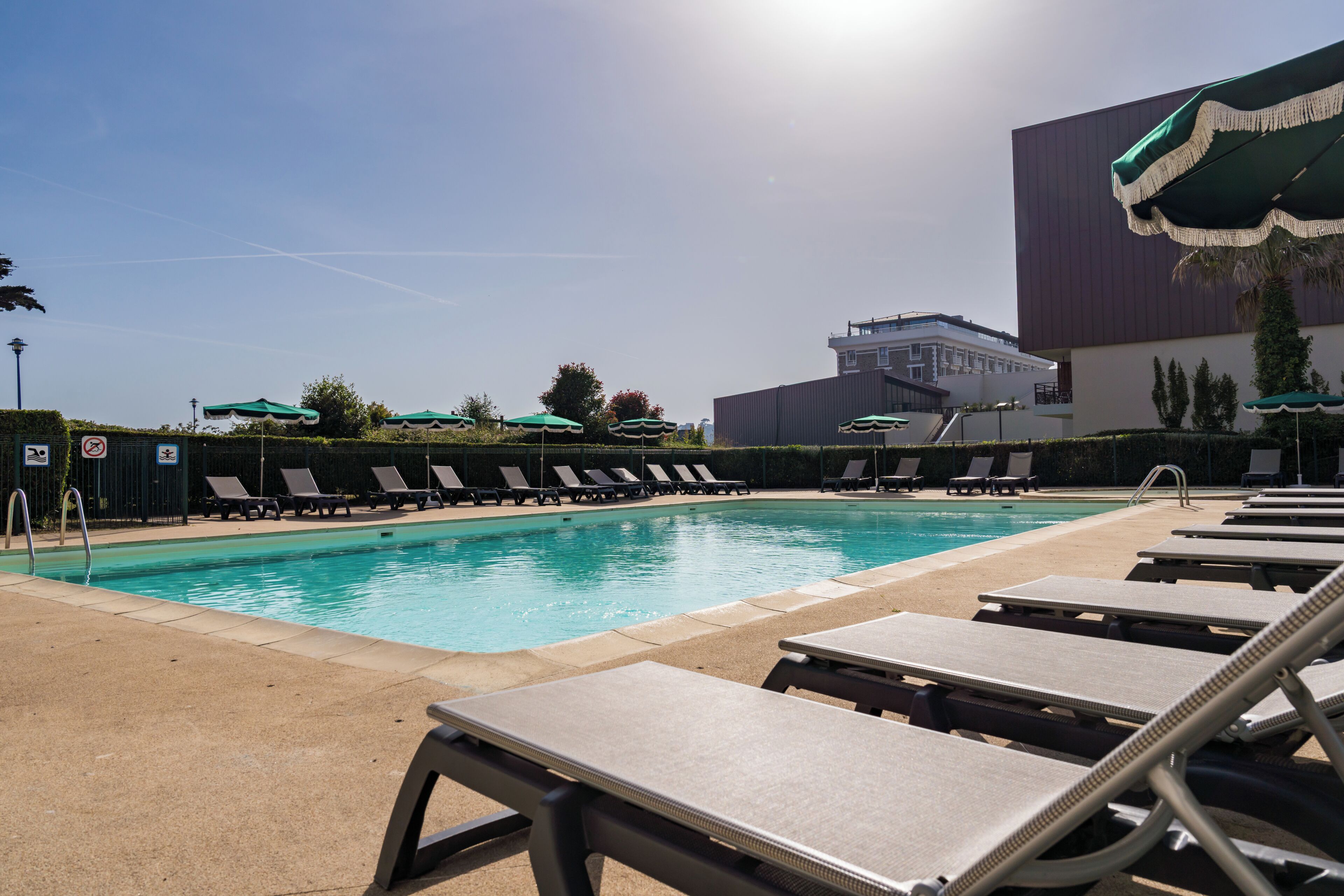 A swimming pool with lounge chairs and umbrellas, a building in the background, and a clear sky.