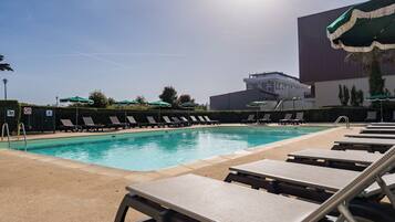 A swimming pool with lounge chairs and umbrellas, a building in the background, and a clear sky.