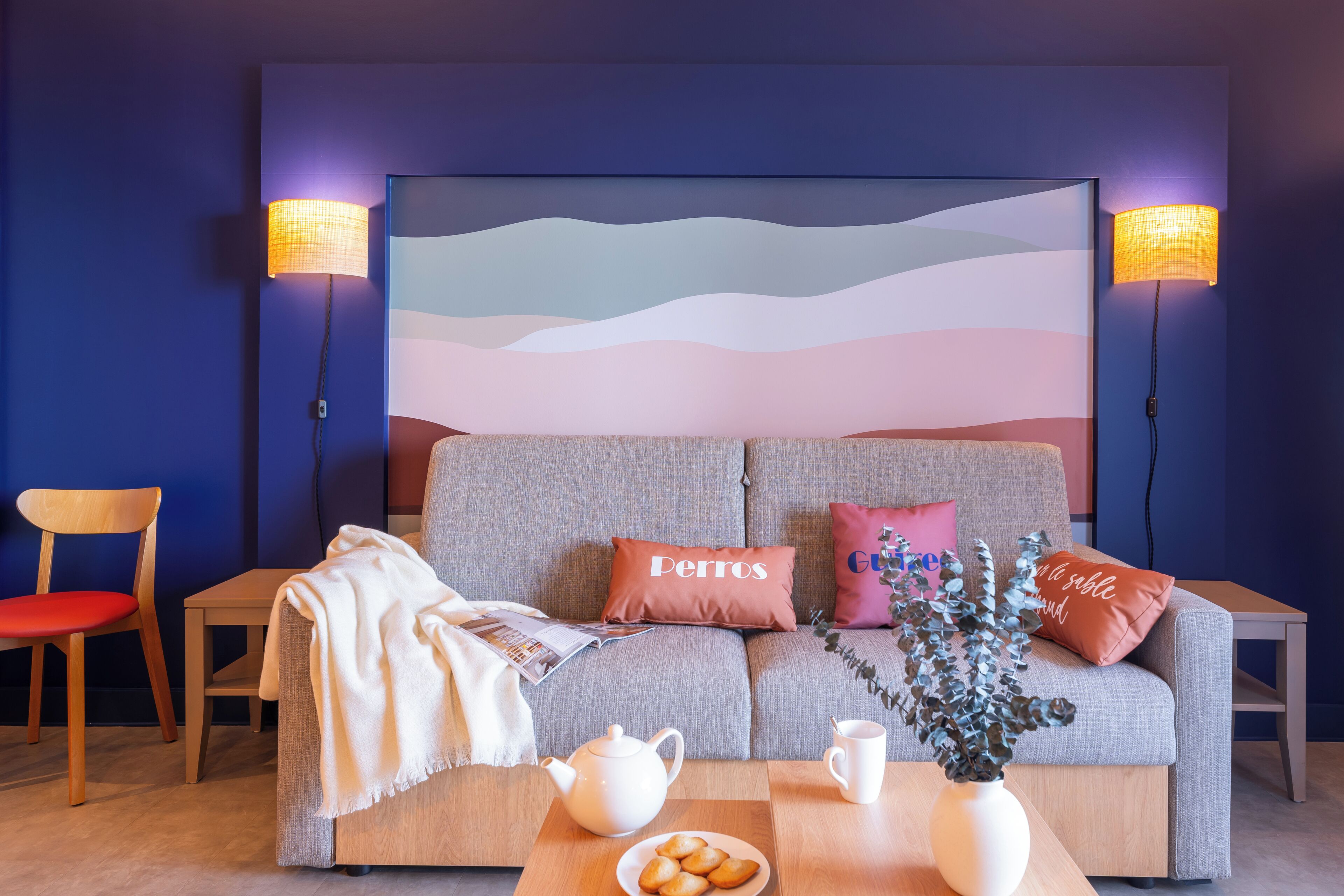 Een moderne woonkamer met een bank, twee stoelen, een bijzettafeltje met een theepot, een bord koekjes en een kunstwerk aan de muur.