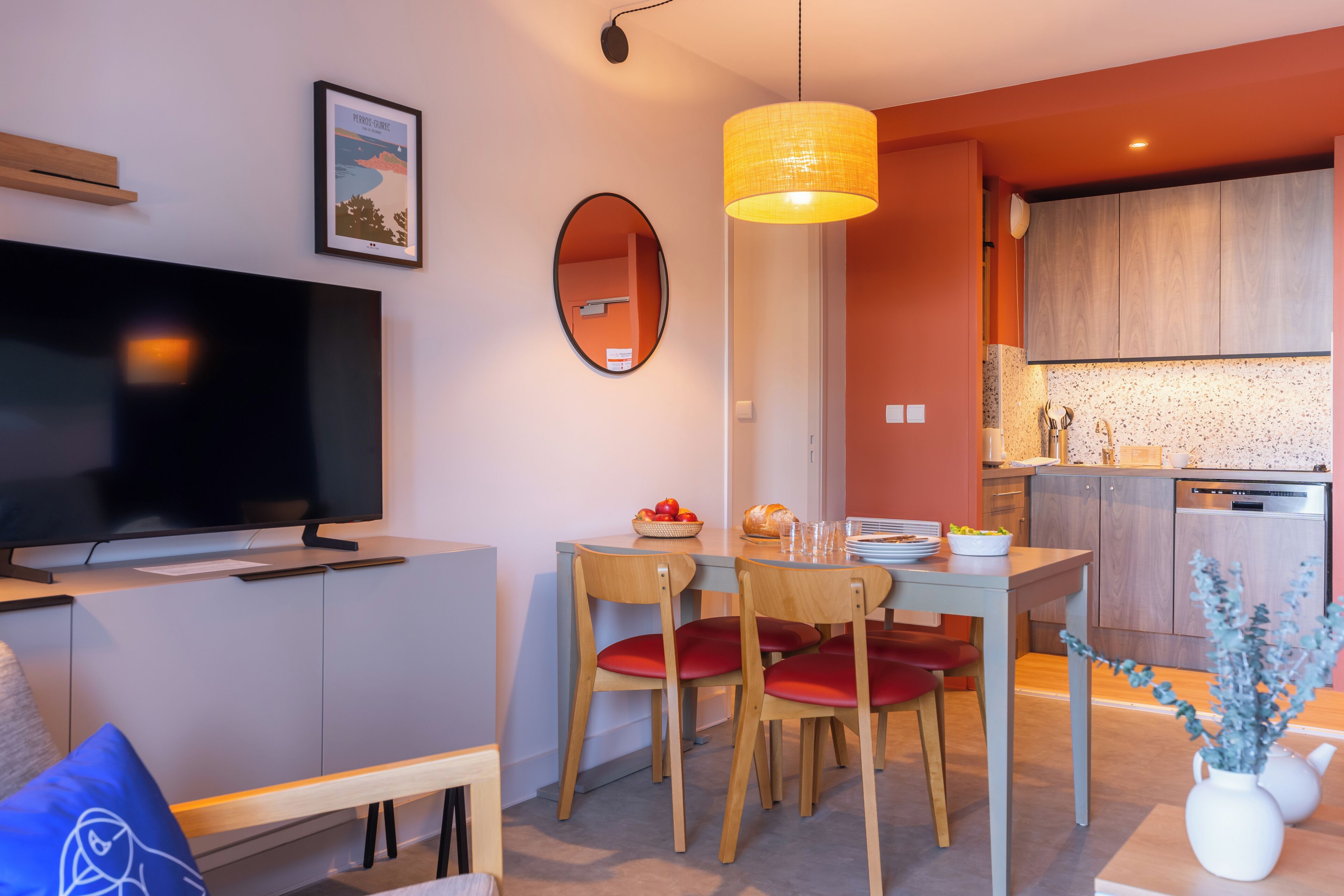A modern living room with a flat-screen TV, a dining table with chairs, a kitchen area with wooden cabinets, and a hanging pendant light.