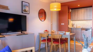 A modern living room with a flat-screen TV, a dining table with chairs, a kitchen area with wooden cabinets, and a hanging pendant light.