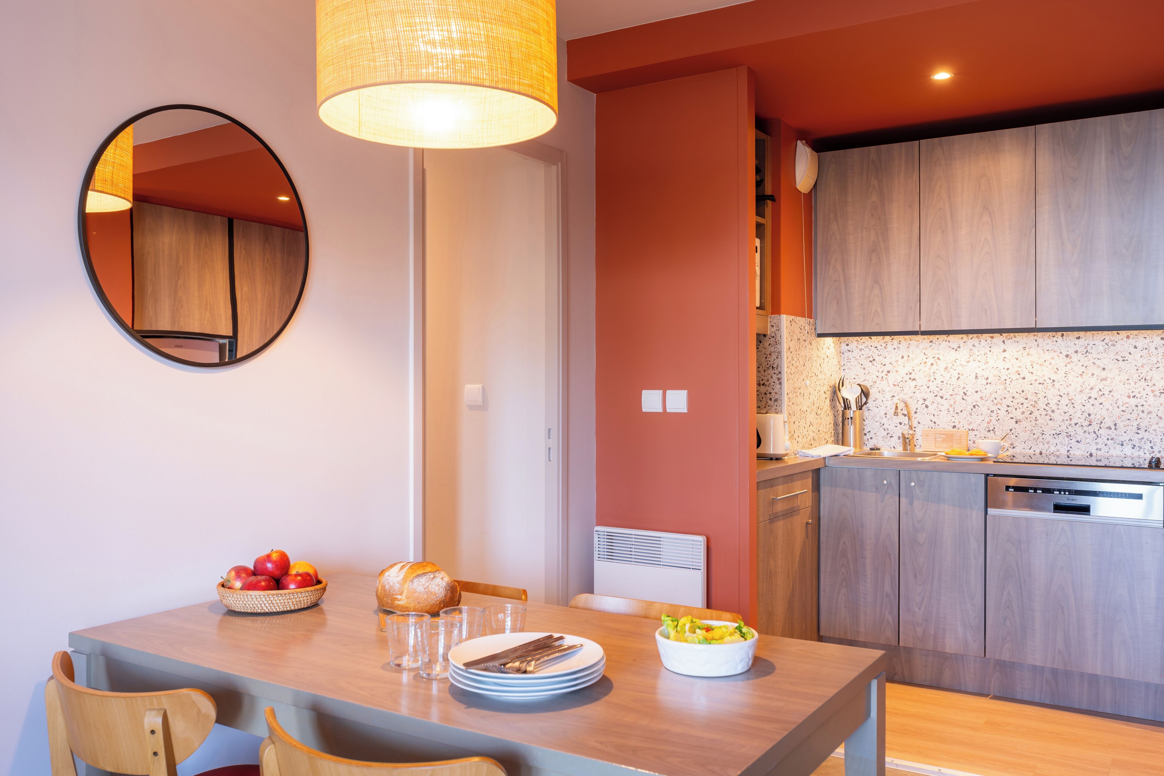Un coin repas agrémenté d’un miroir rond, d’une table mise avec des assiettes, des verres et un panier de fruits.