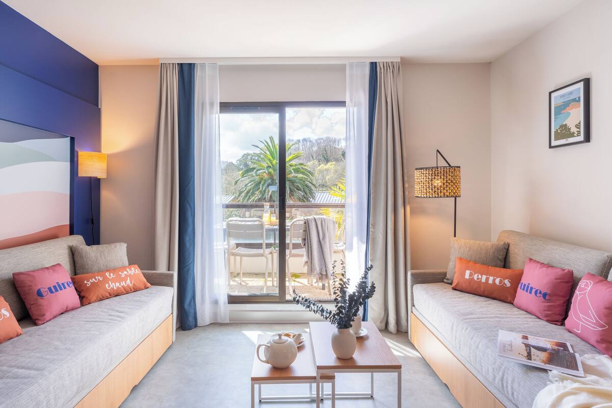 A modern hotel room with two sofas, a table with a vase and a teapot, and a view of the outdoors.