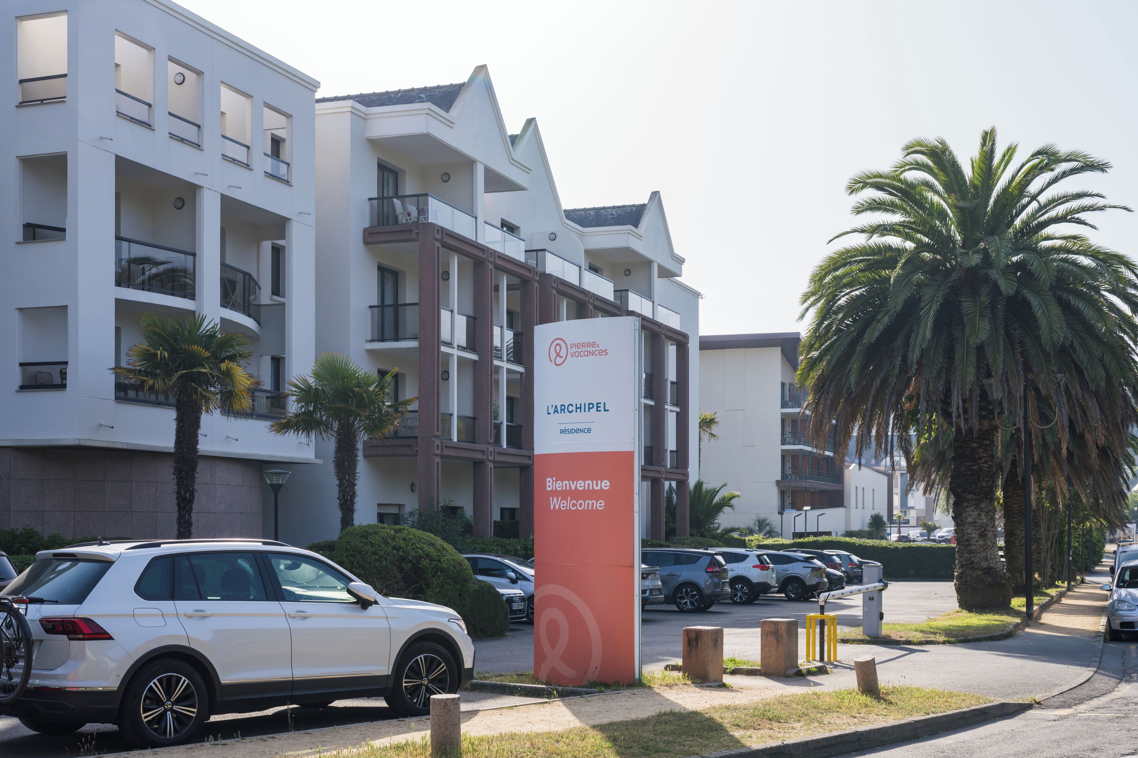 A multi-story hotel with balconies, a signboard with 'Bienvenue Welcome,' and parked cars.