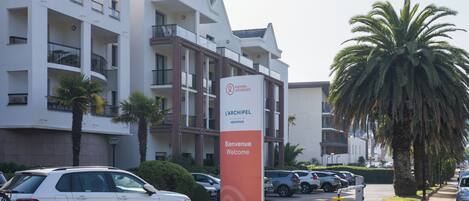 A multi-story hotel with balconies, a signboard with 'Bienvenue Welcome,' and parked cars.