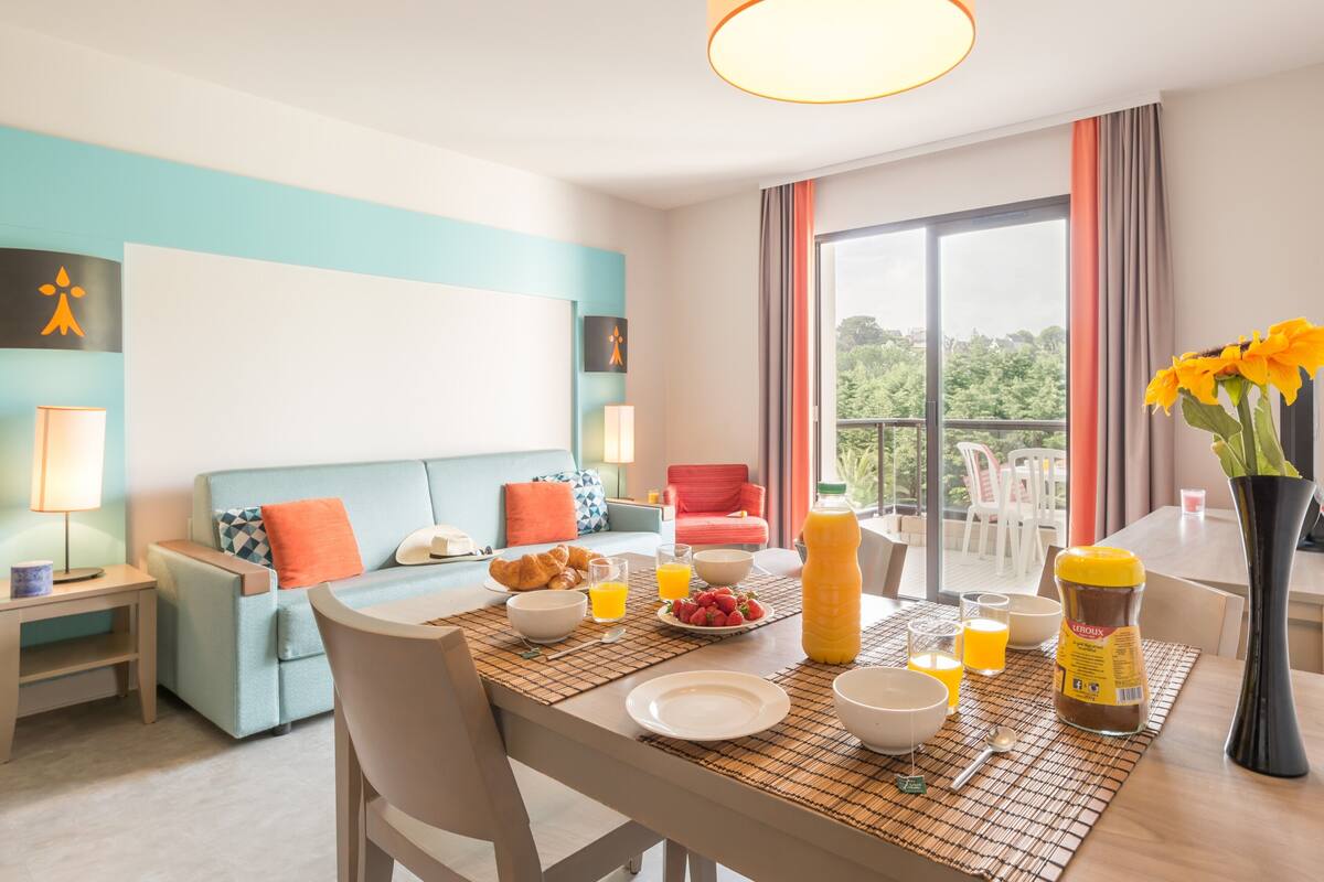 A modern hotel room with a dining area, breakfast setup, and a balcony with a view.