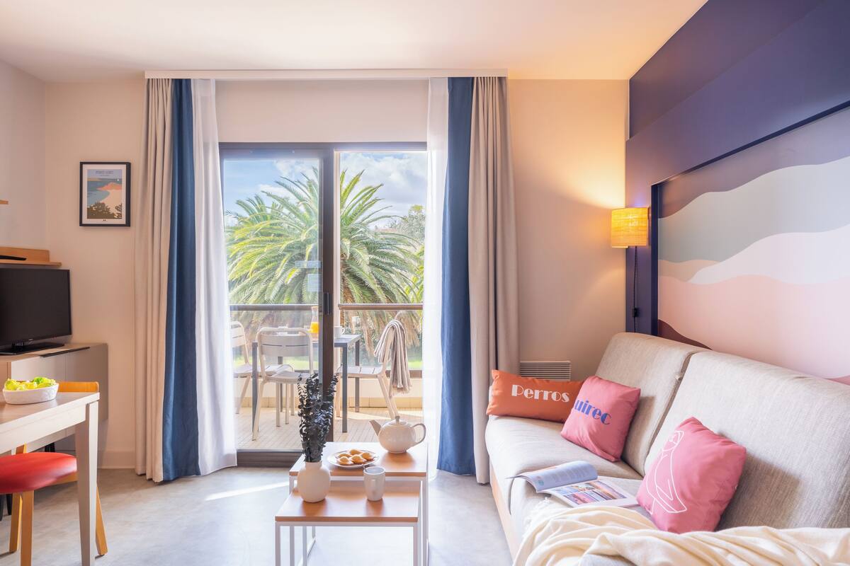 A modern hotel room with a balcony, a sofa with pink pillows, a television, and a dining area with a table and chairs.