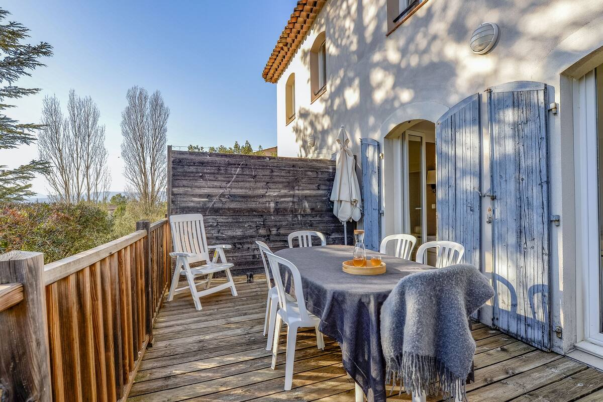 Une terrasse en bois avec une table et des chaises, un espace extérieur aménagé et couvert, et un bâtiment avec une porte bleue.