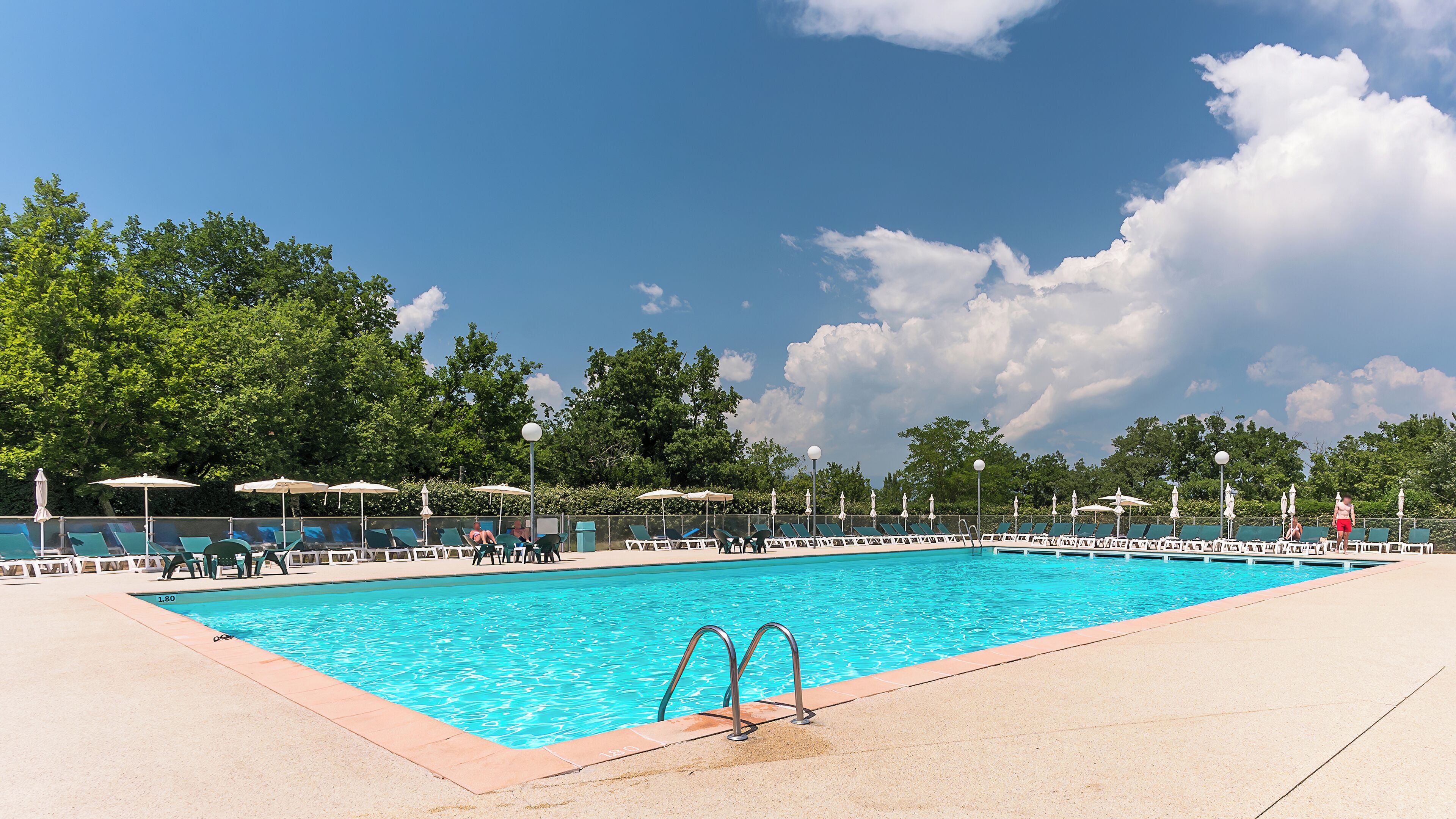 Une piscine rectangulaire à l’eau d’un bleu limpide, entourée d’une terrasse en béton et bordée de nombreuses chaises longues et de parasols.