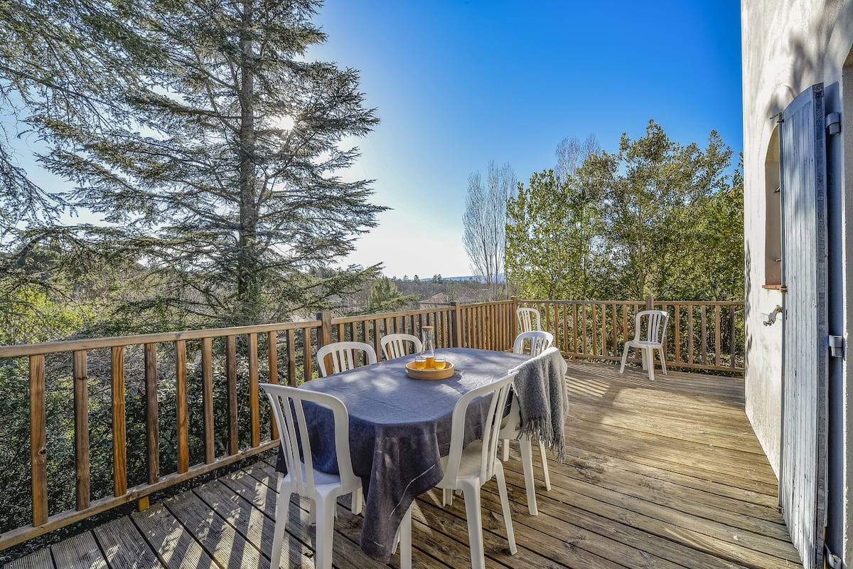 Une terrasse en bois avec une table dressée pour le repas, entourée de chaises blanches, donnant sur une zone boisée.