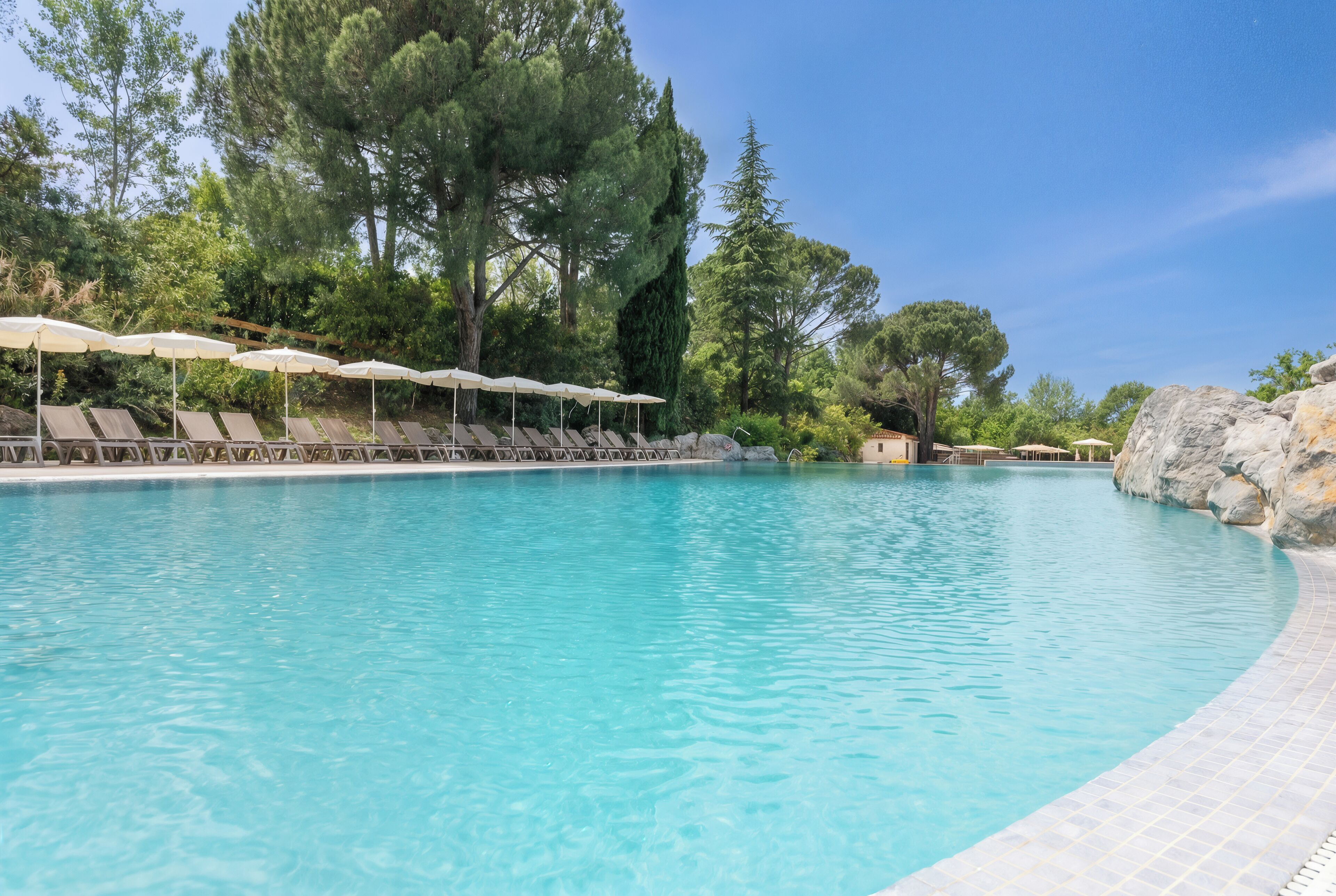 A swimming pool with lounge chairs and umbrellas, surrounded by trees and rocks.