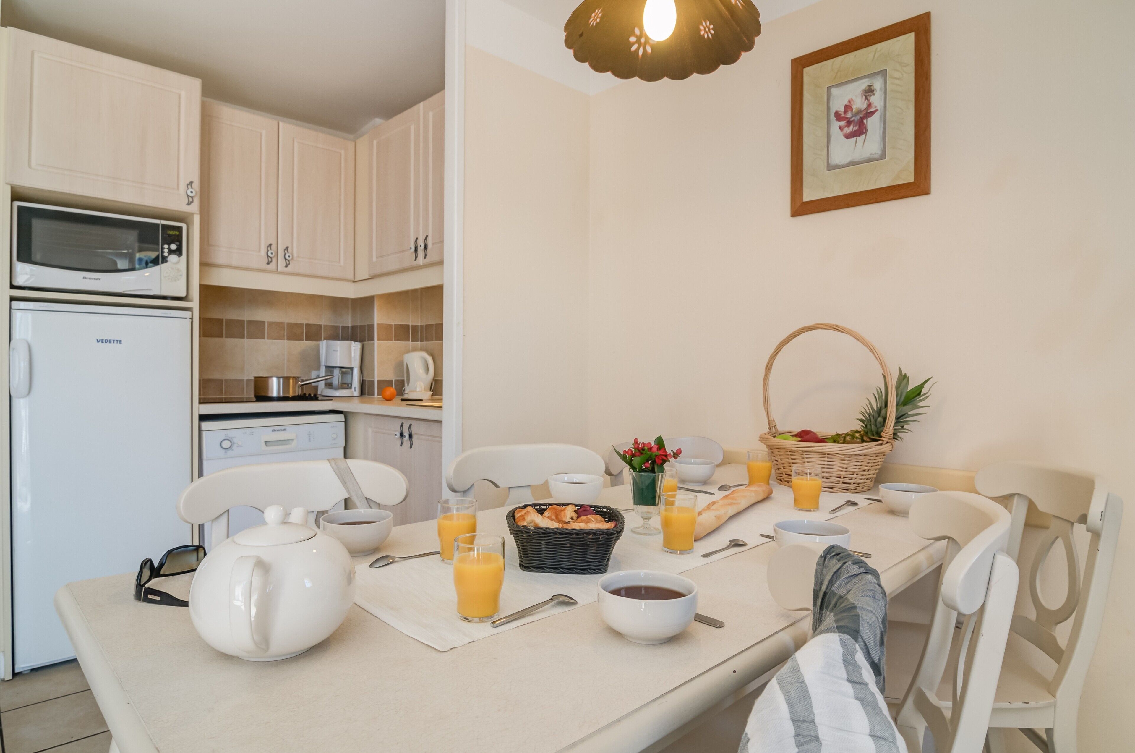 A kitchen with white cabinets, a microwave, a coffee maker, and a dining table set with a basket of fruit, bowls, and glasses of orange juice.