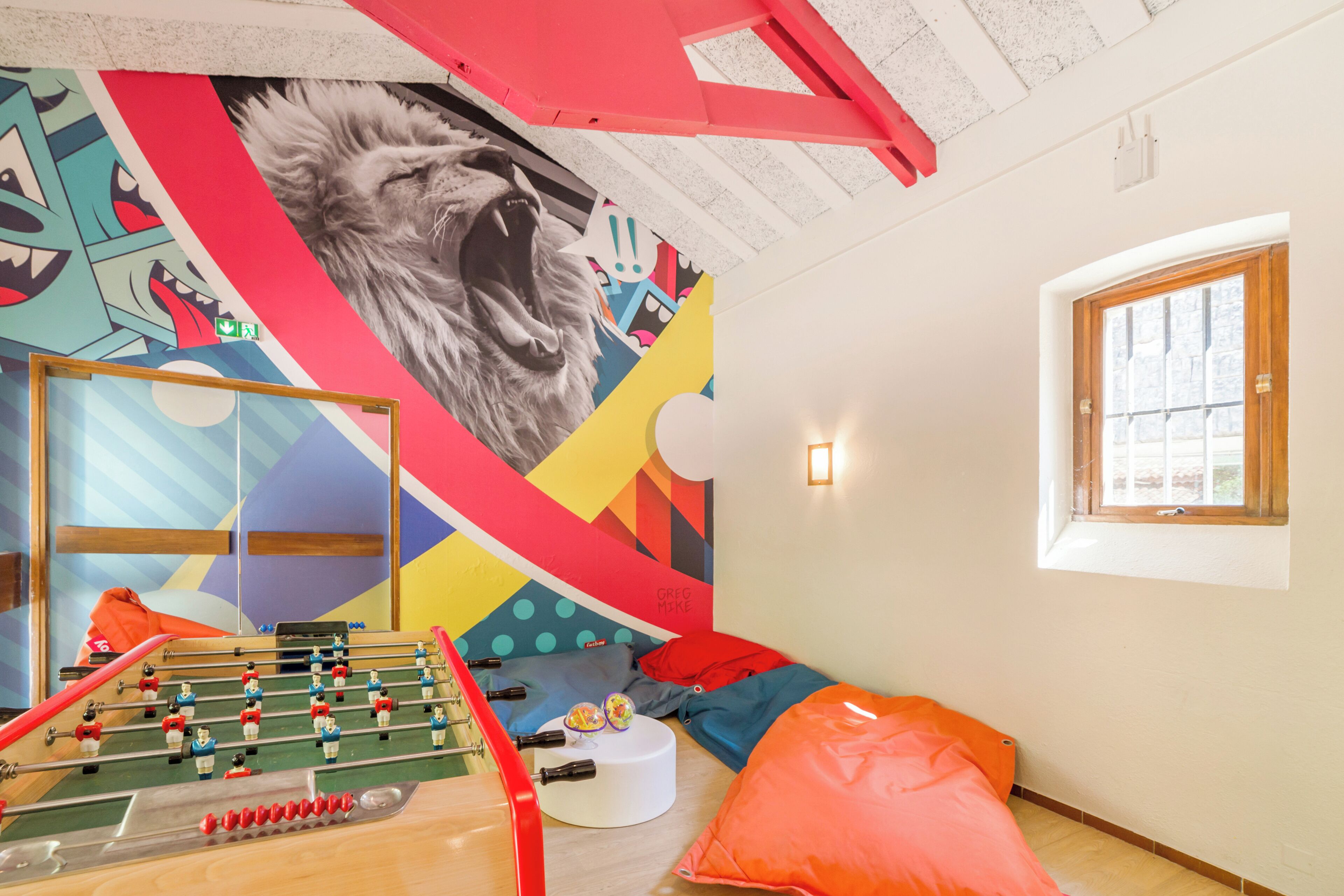 A room with a foosball table, colorful wall art, a window with a view, and a bed with pillows.