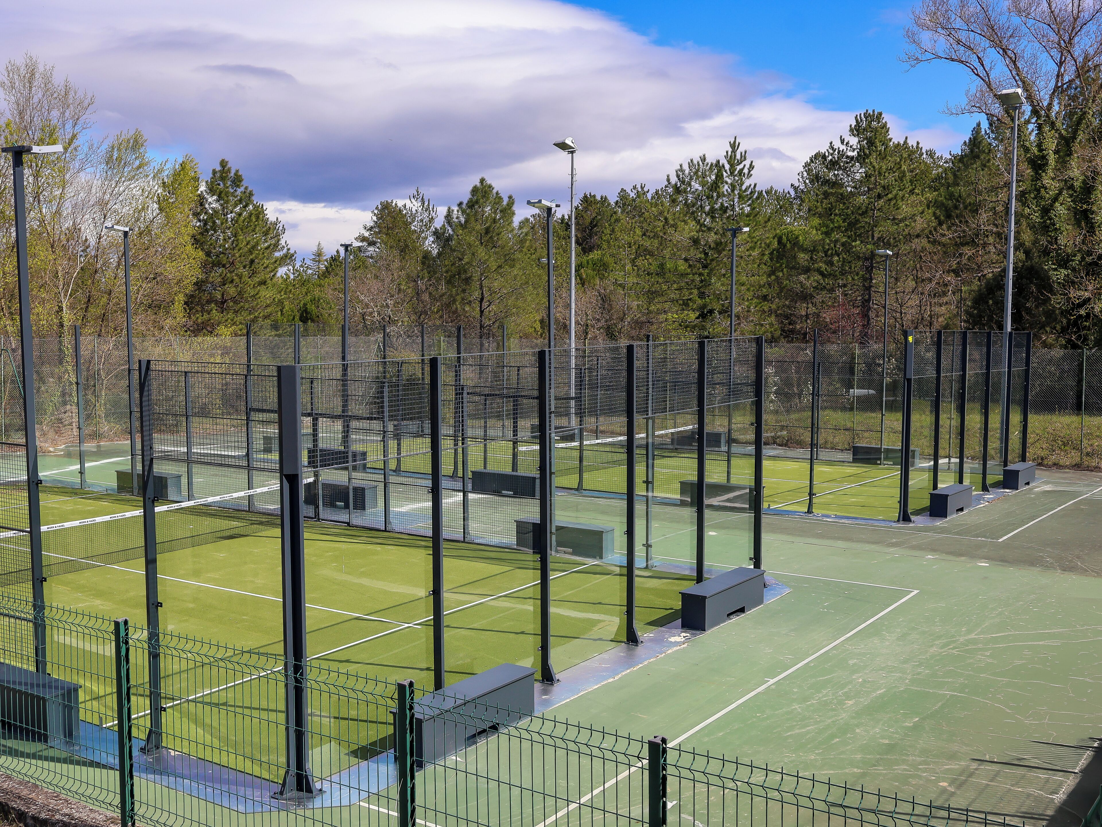 Un court de tennis avec un filet, entouré d’une clôture, et des arbres en arrière-plan.