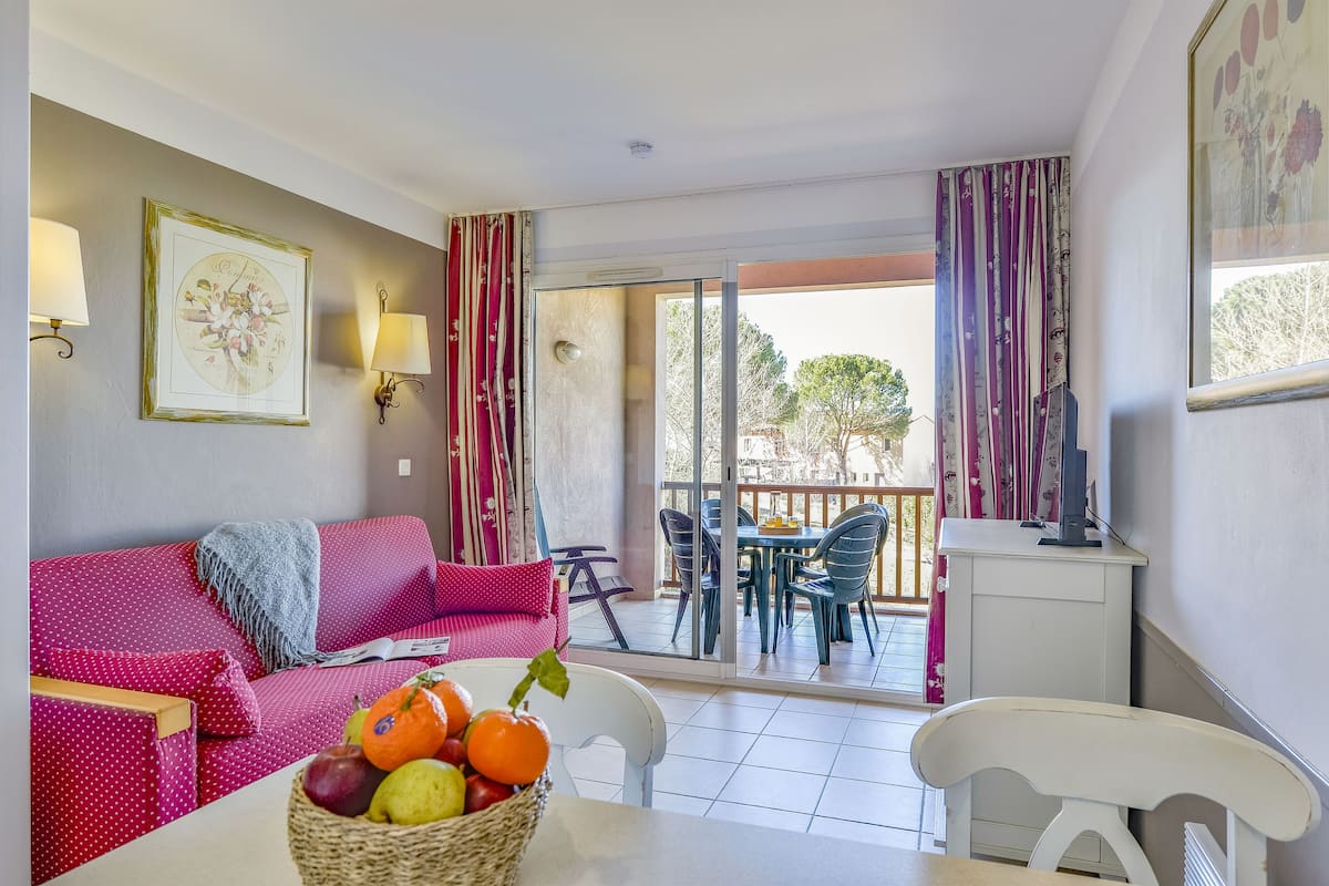 Un salon chaleureux avec un canapé rouge à pois, une table blanche sur laquelle est posé un panier de fruits, et un balcon agrémenté de mobilier d’extérieur.