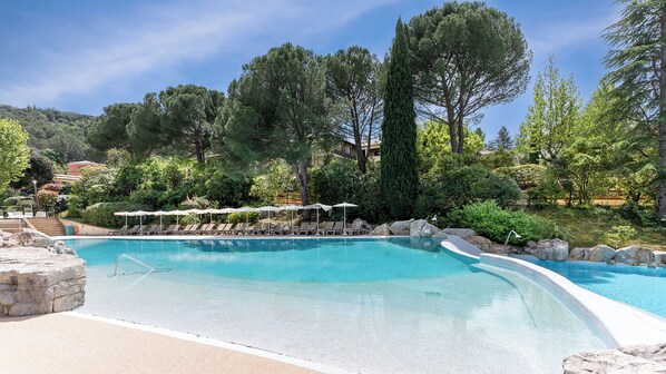 A pool with lounge chairs and umbrellas, surrounded by trees and a hillside.