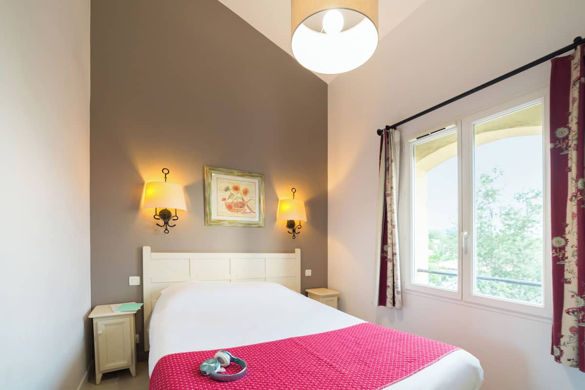 Un lit bien fait, recouvert d’une couverture rouge, deux lampes de chevet, un tableau encadré au mur et une fenêtre avec des rideaux.