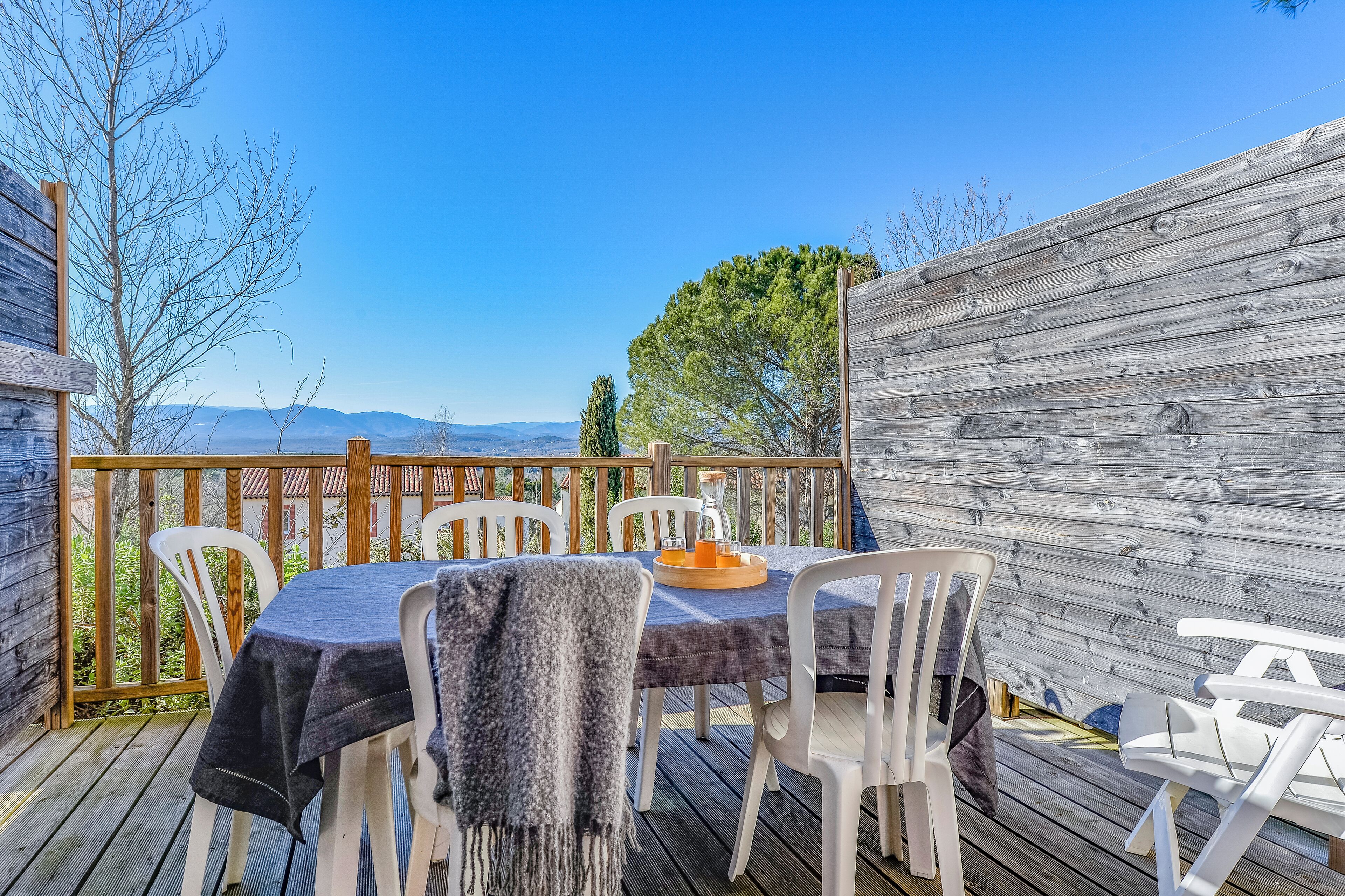 An outdoor patio with a table set for two, surrounded by white chairs, overlooking a scenic view of mountains and trees.