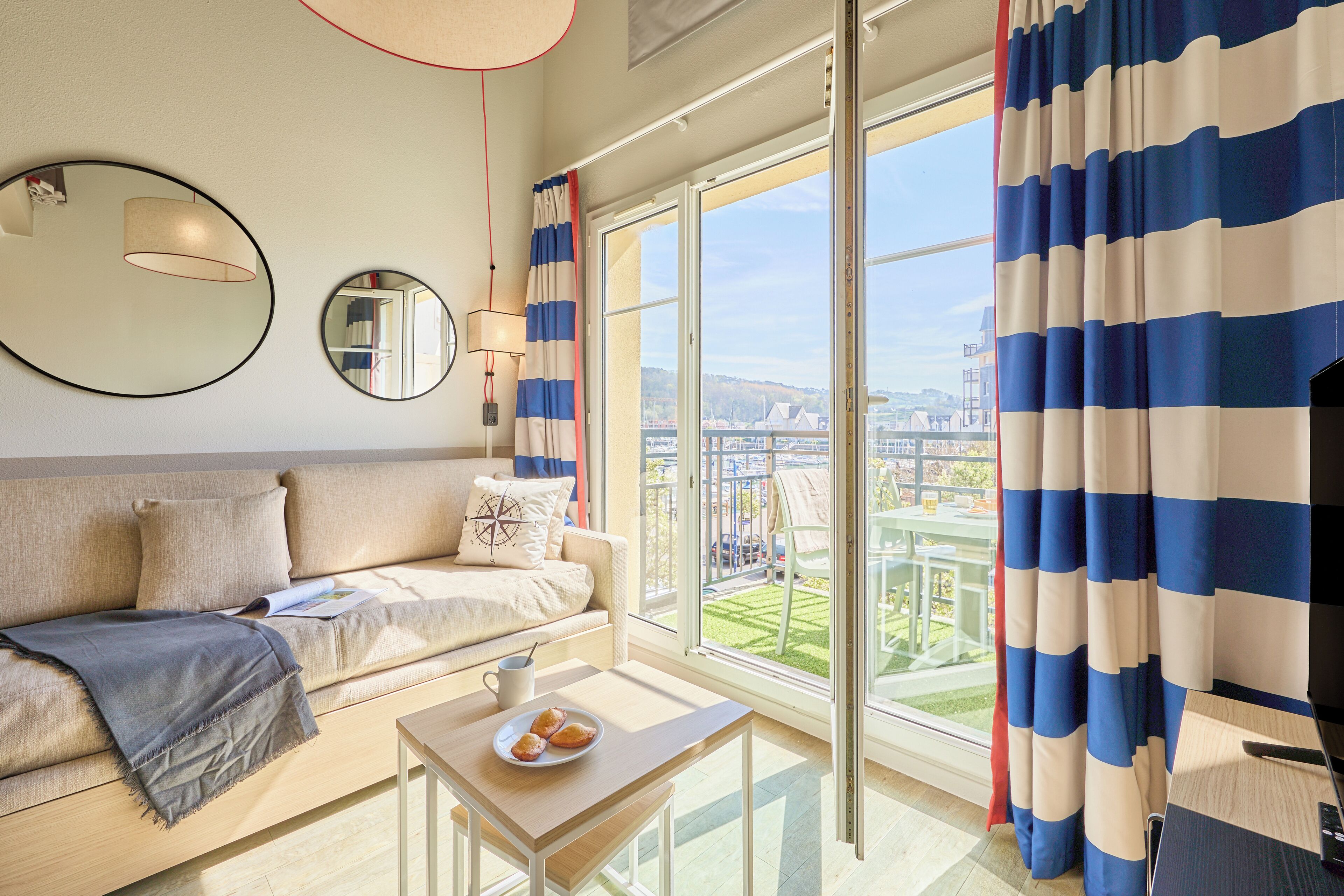 Une chambre d’hôtel moderne avec un canapé, une table sur laquelle se trouve une assiette de pâtisseries, une lampe et de grandes fenêtres avec des rideaux.