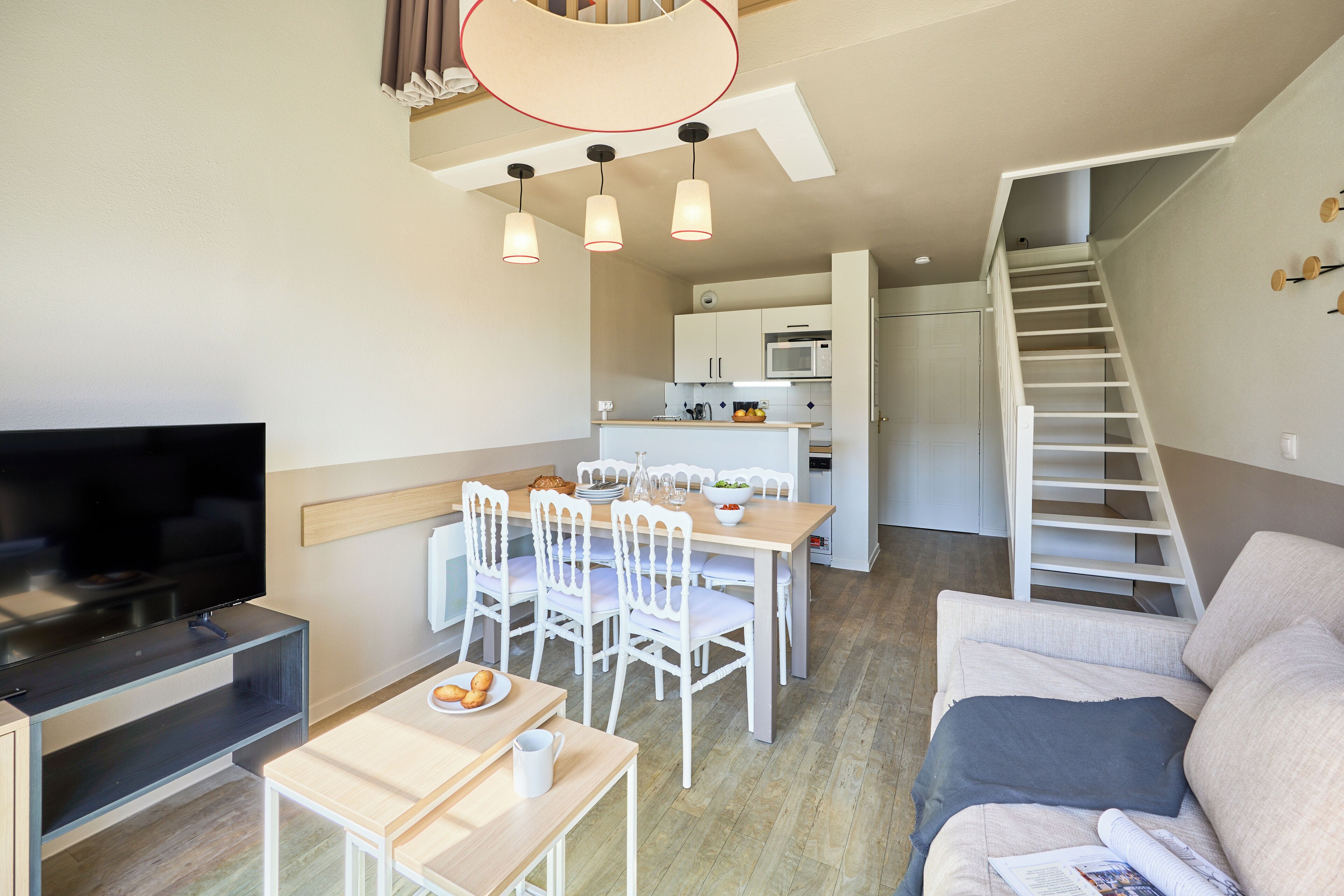 A modern living room with a flat-screen TV, a dining area with white chairs and a wooden table, and a staircase leading to an upper level.