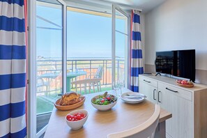 A balcony with a clear view of the sea, a dining table with a salad, bread, and a bowl of cherries, and a TV on the wall. - Pierre & Vacances Résidence Port Guillaume (Dives-sur-Mer)