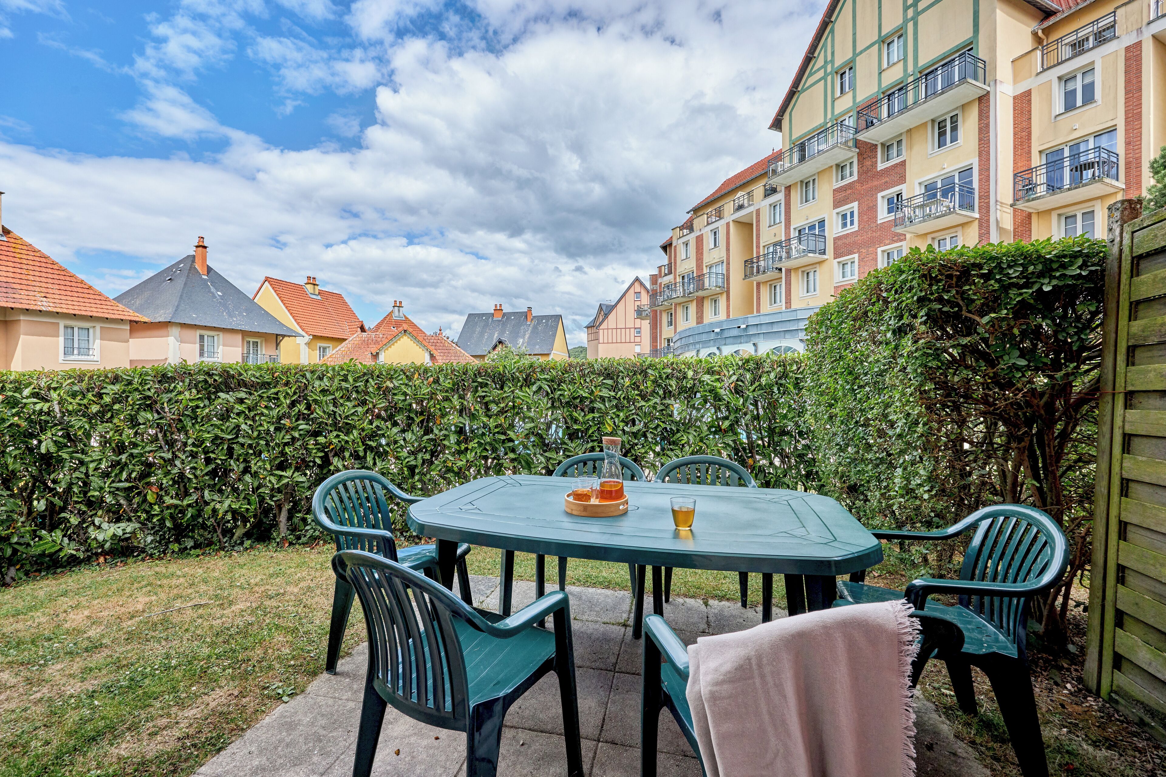 A patio with a table and chairs, surrounded by a hedge and buildings in the background.