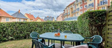 A patio with a table and chairs, surrounded by a hedge and buildings in the background.