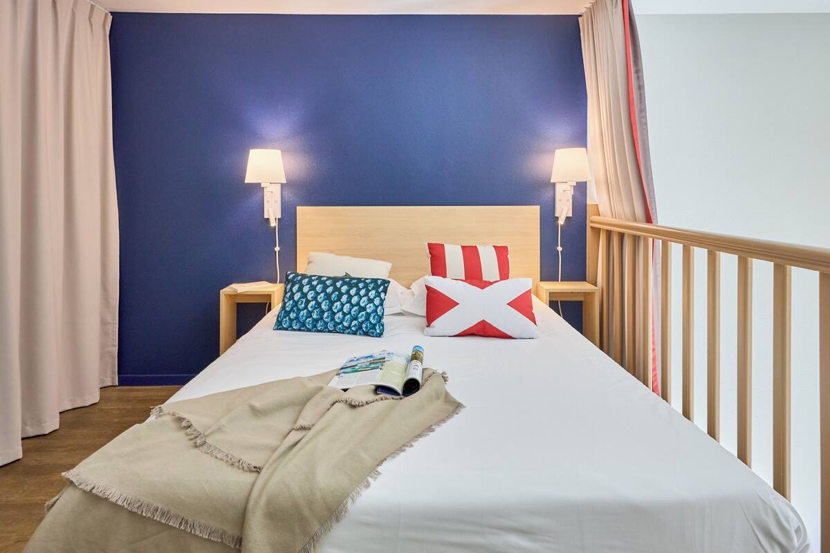 Ein ordentlich bezogenes Bett mit einem weiß-roten Kopfkissen, einem blauen Kopfkissen und einer beige farbenen Tagesdecke. Zwei Nachttischlampen und ein Holzkopfbrett sind ebenfalls zu sehen.