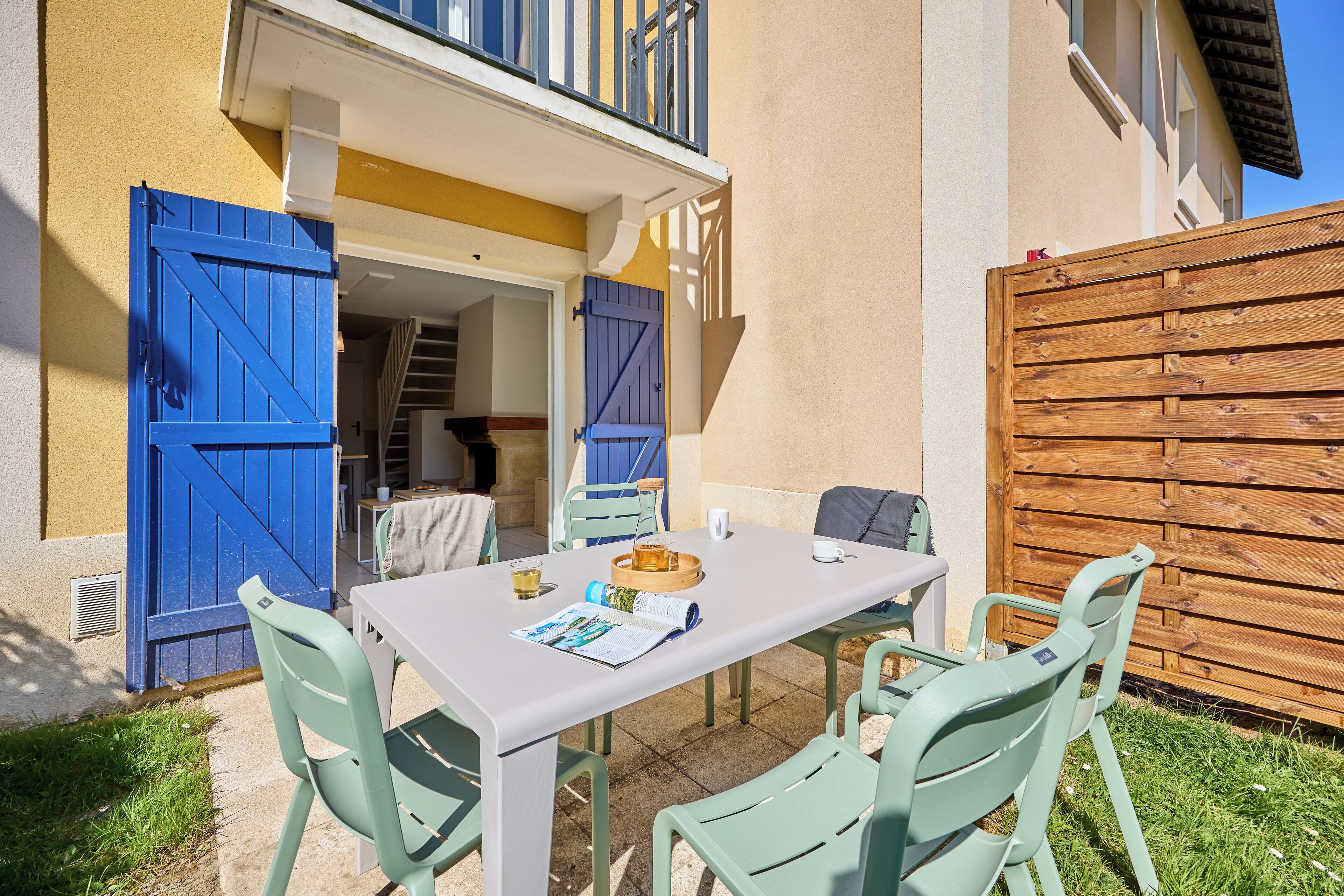 Een terras met een tafel en stoelen, een houten hek en een gebouw met blauwe luiken.