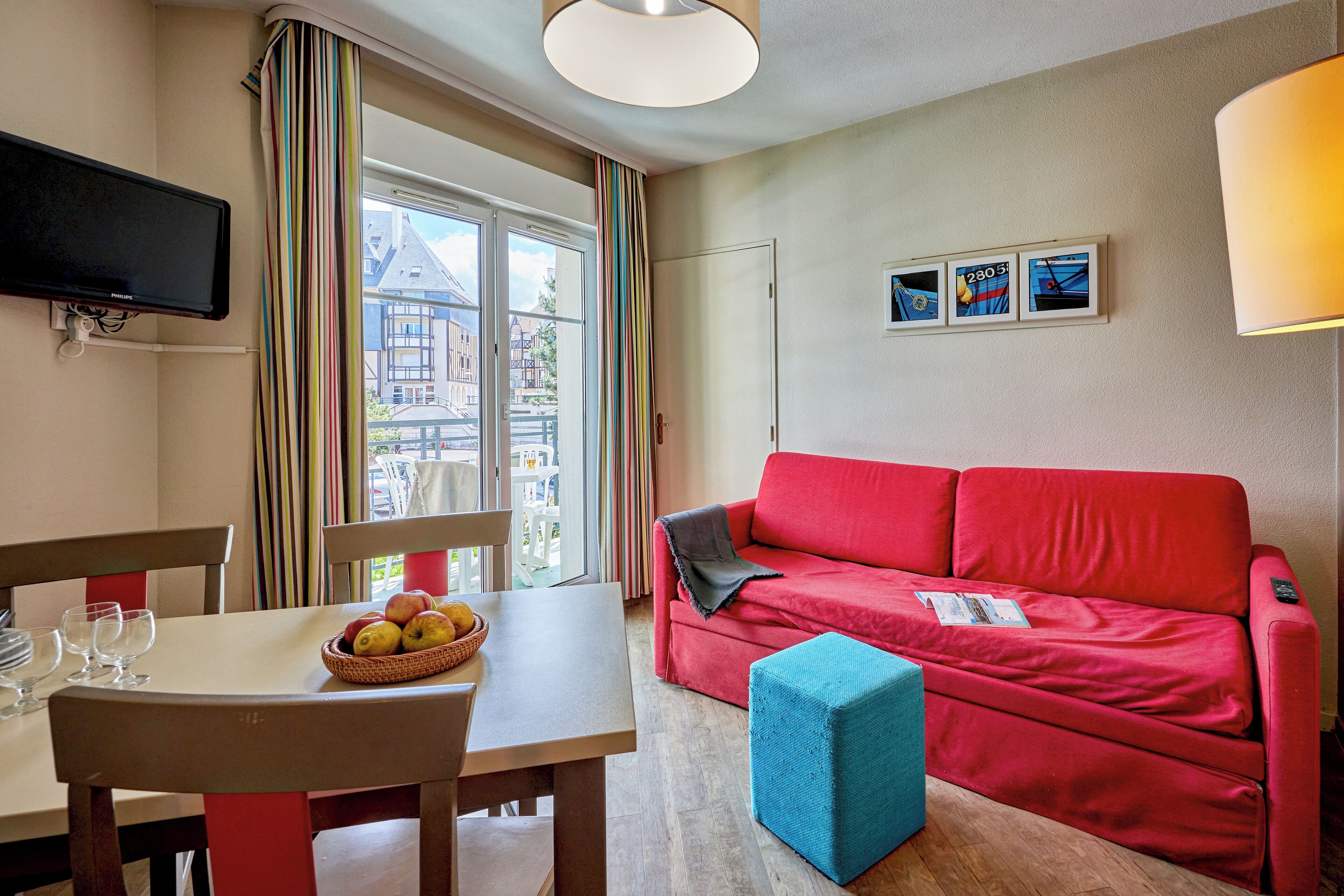 A cozy living room with a red sofa, a small blue ottoman, a dining table with chairs, and a flat-screen TV mounted on the wall.