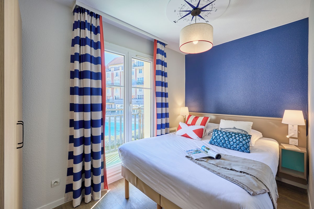 Ein Hotelzimmer mit einem großen Bett, einem Nachttisch mit Lampe, einem Deckenventilator und Blick auf Gebäude durch das Fenster.