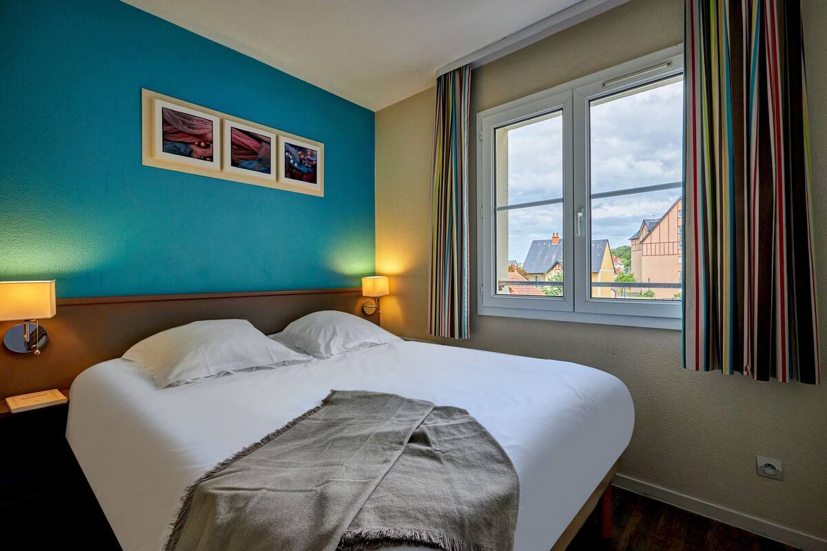 Ein Hotelzimmer mit einem großen Bett, einem Nachttisch mit Lampe und einem Fenster mit Vorhängen.