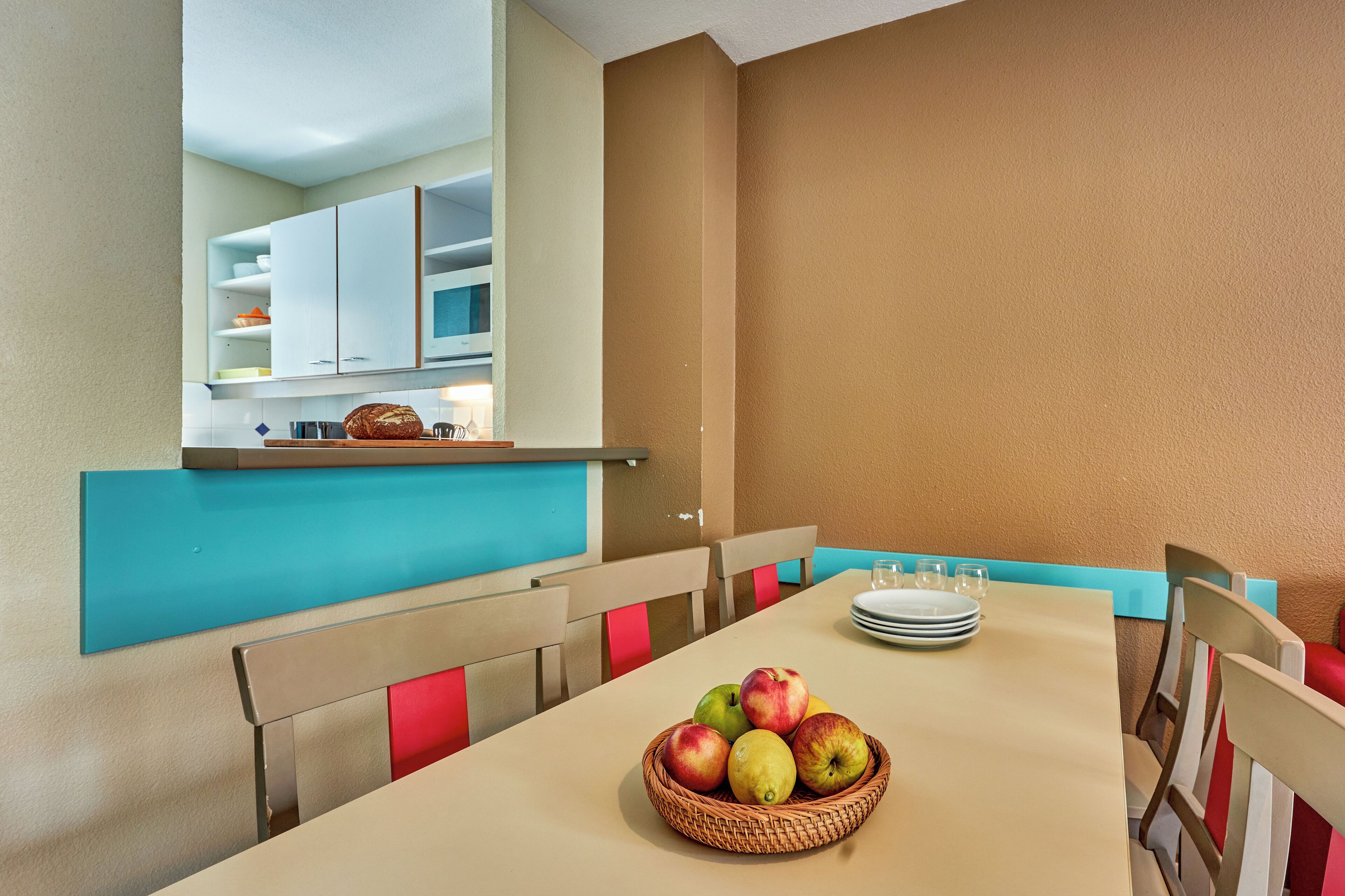 Un coin repas avec une table dressée pour quatre personnes, un panier de fruits et une vue sur une cuisine dotée de meubles blancs et d’un plan de travail bleu.