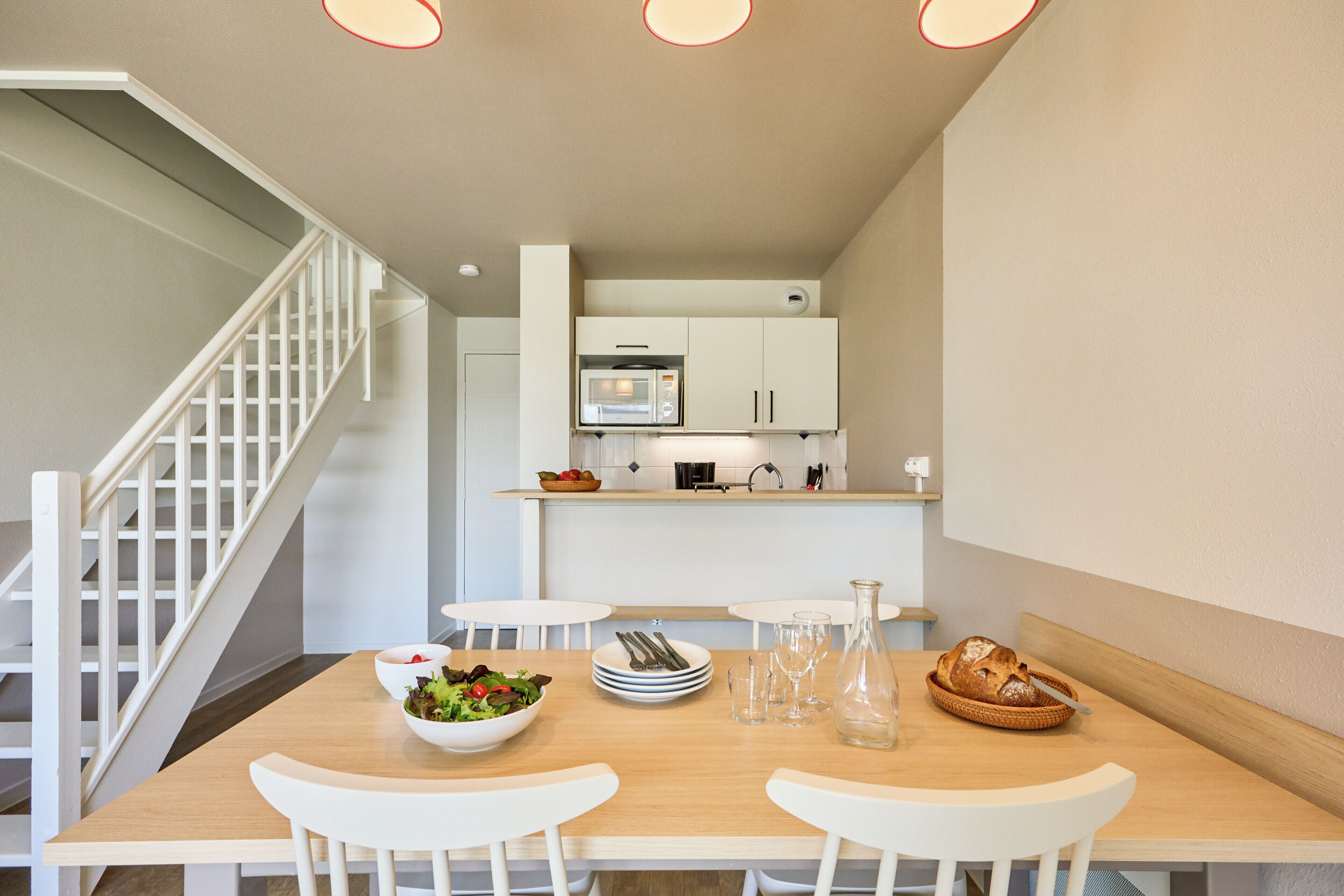 Een eethoek met een houten tafel, witte stoelen, een saladekom, borden, bestek, een brood en een glazen fles.